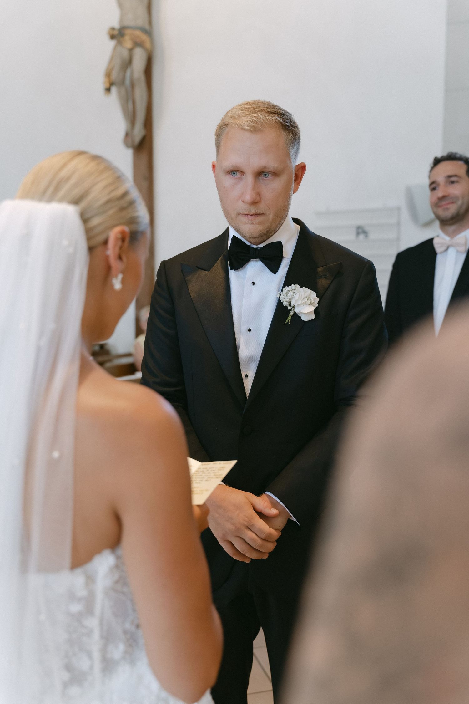 Groom in a black tuxedo with bow tie and white boutonniere listens intently to the bride in a veil during a church wedding ceremony.
