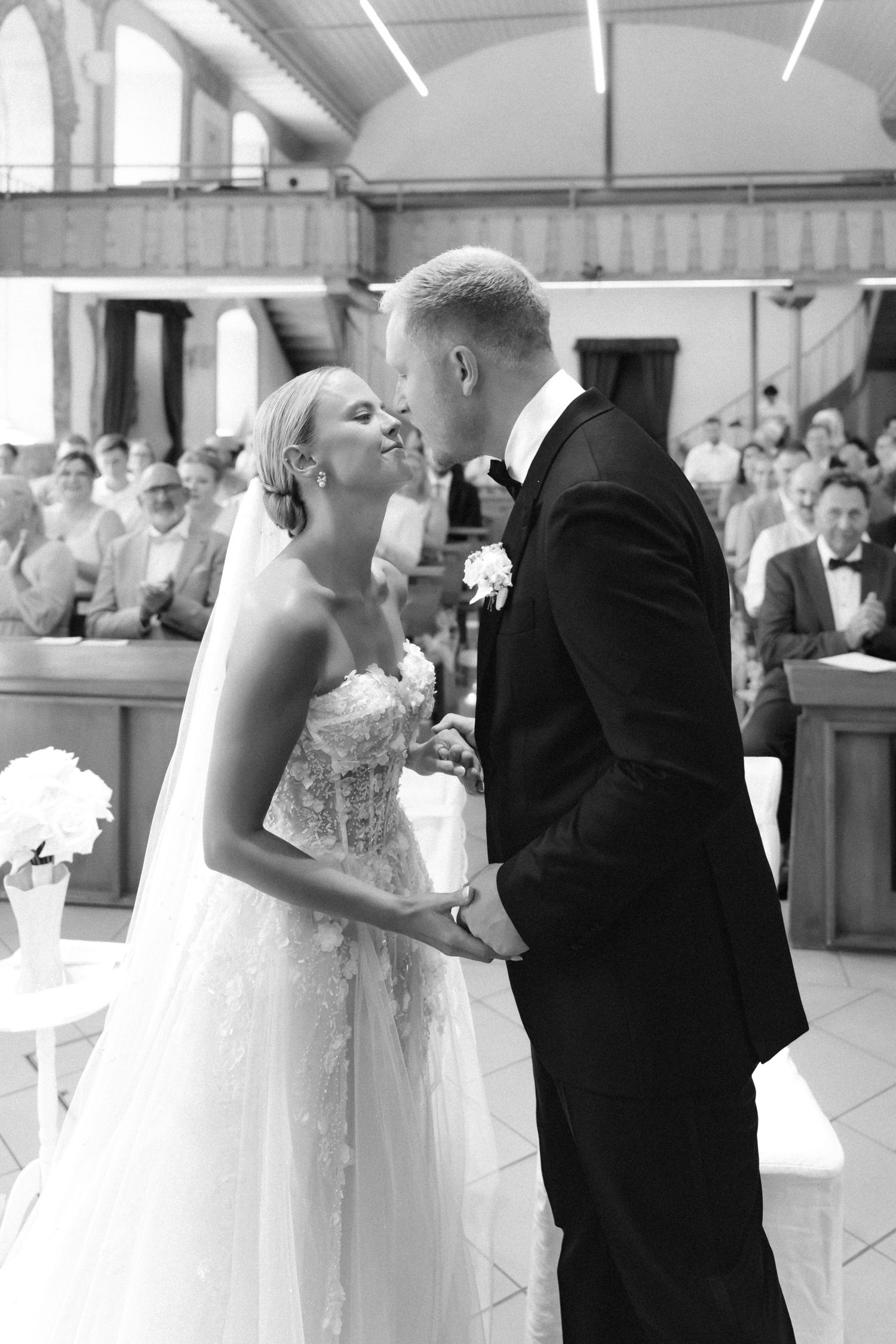 Black and white photo of a bride in a lace gown and groom in a suit sharing a wedding kiss in front of seated guests.