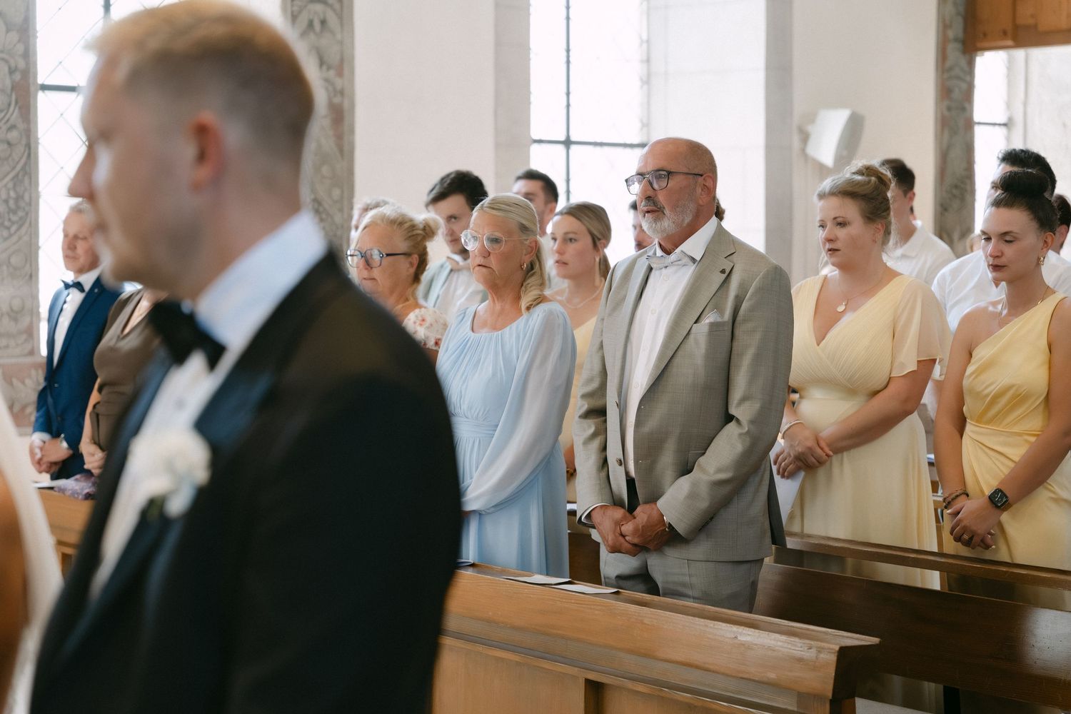 Groom in a black tuxedo with a white boutonniere stands in a church pew as wedding guests in formal attire look on during the ceremony.