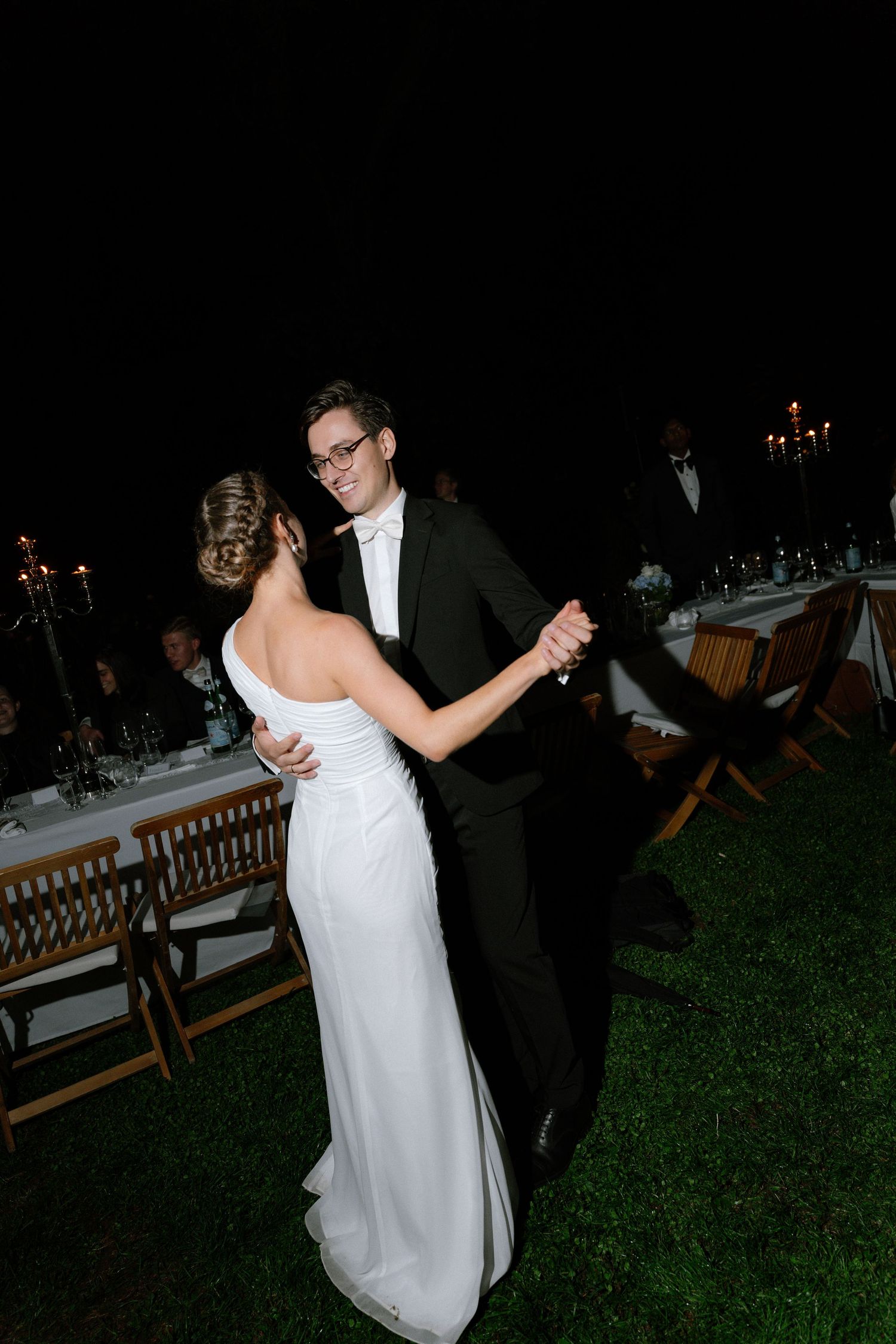 Bride in an elegant white gown dances with a groom in a dark suit and glasses on a lawn at a candlelit outdoor evening wedding reception.
