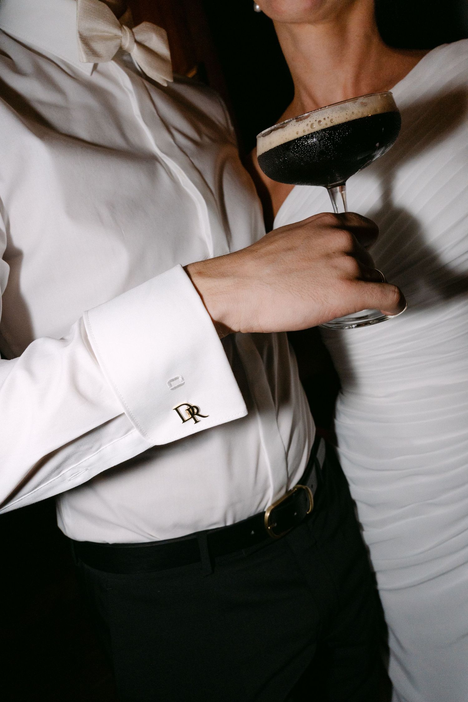 Person in a white dress shirt with bow tie and monogrammed DR cufflink holds an espresso martini beside a woman in a white dress.
