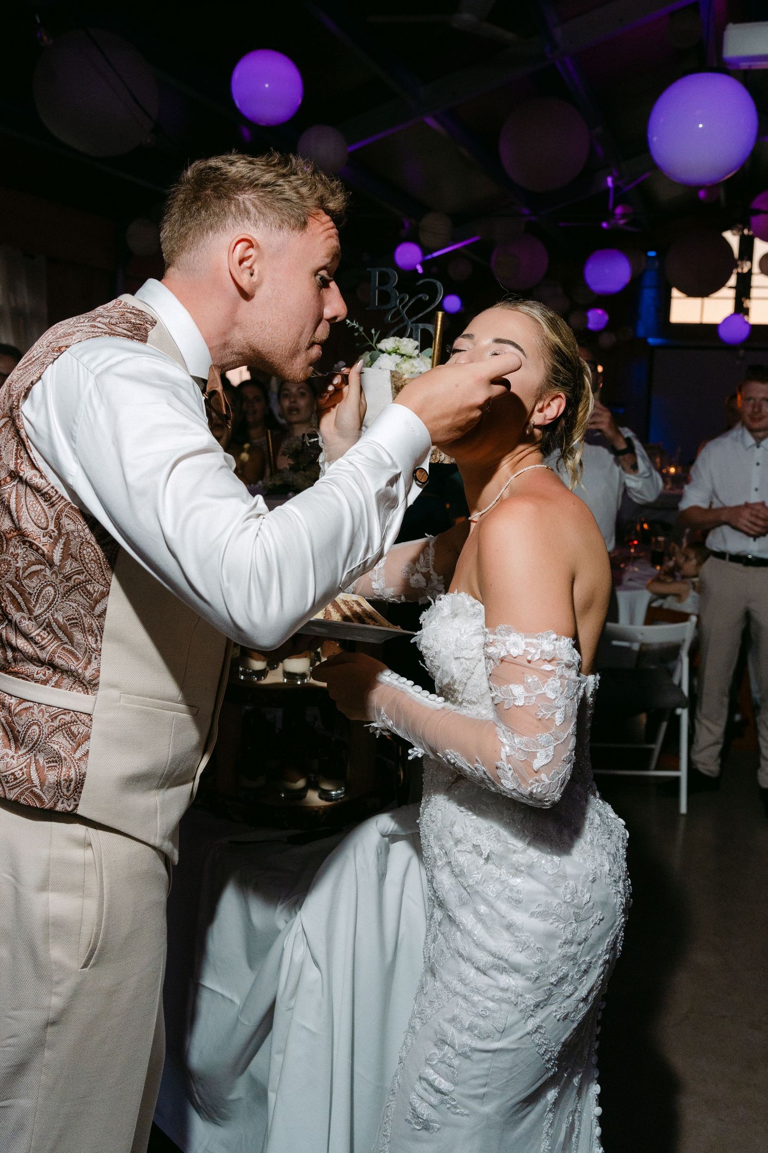 A couple shares a playful cake-feeding moment at their wedding reception with purple balloon decor in the background.