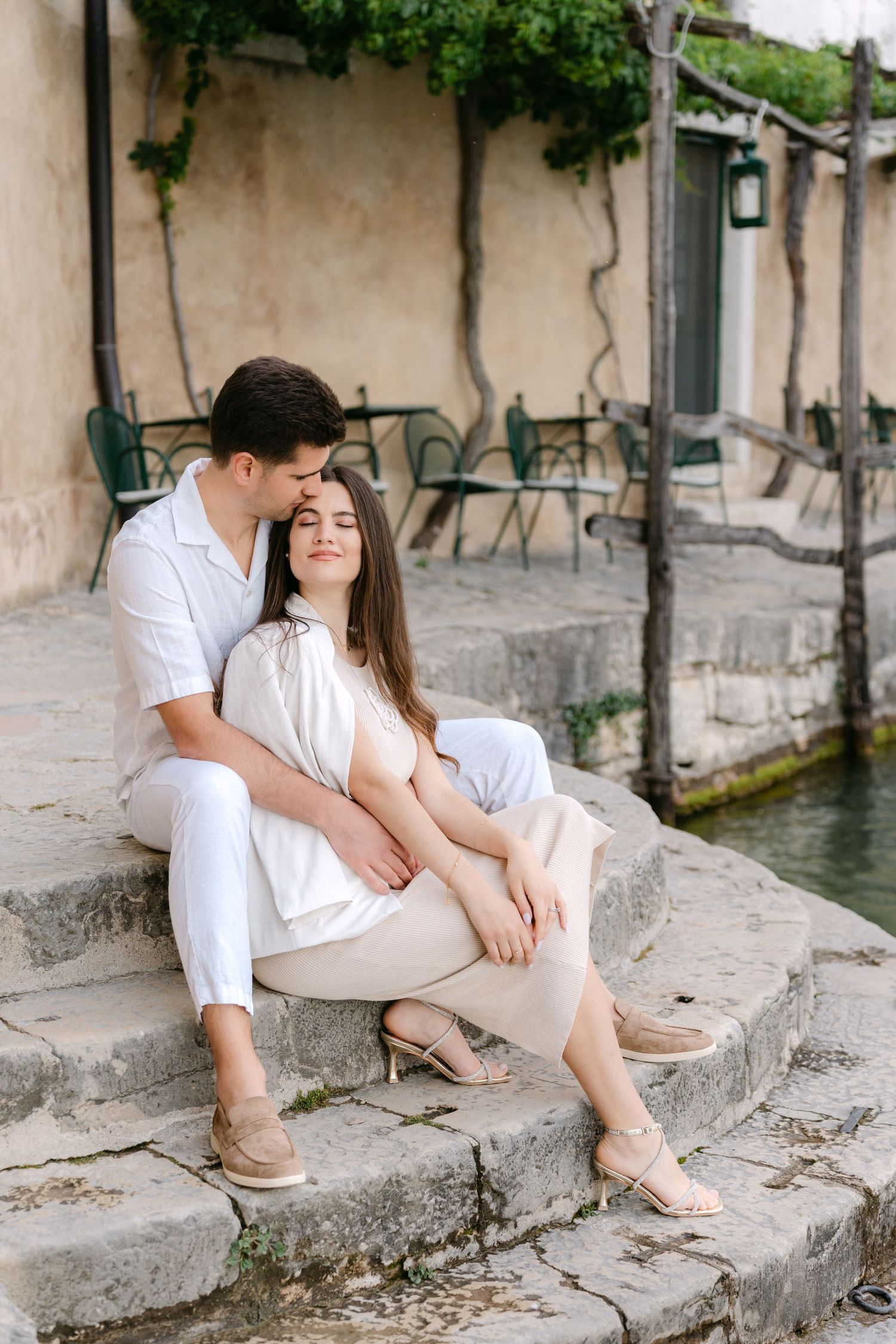 A romantic couple in white outfits sits together on stone steps near a waterway in a European-style setting.