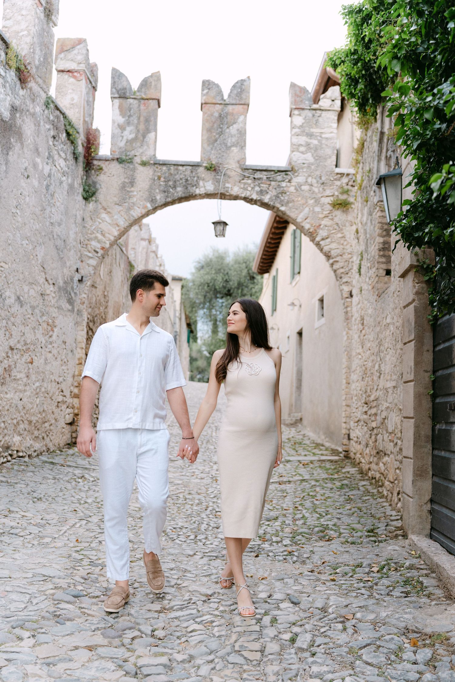 A couple in white outfits walking hand in hand through a charming cobblestone street with ancient stone walls.