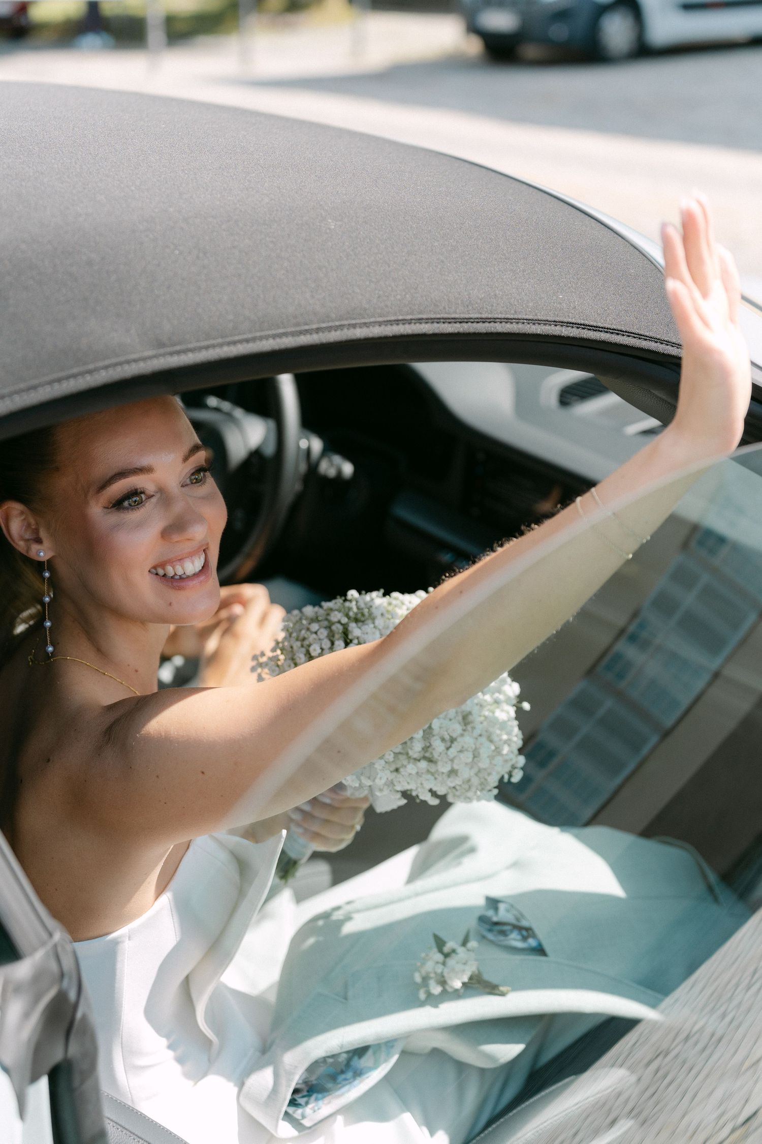 A bride in a white wedding dress waves from inside a car window with floral decorations. Schloss Solitude
