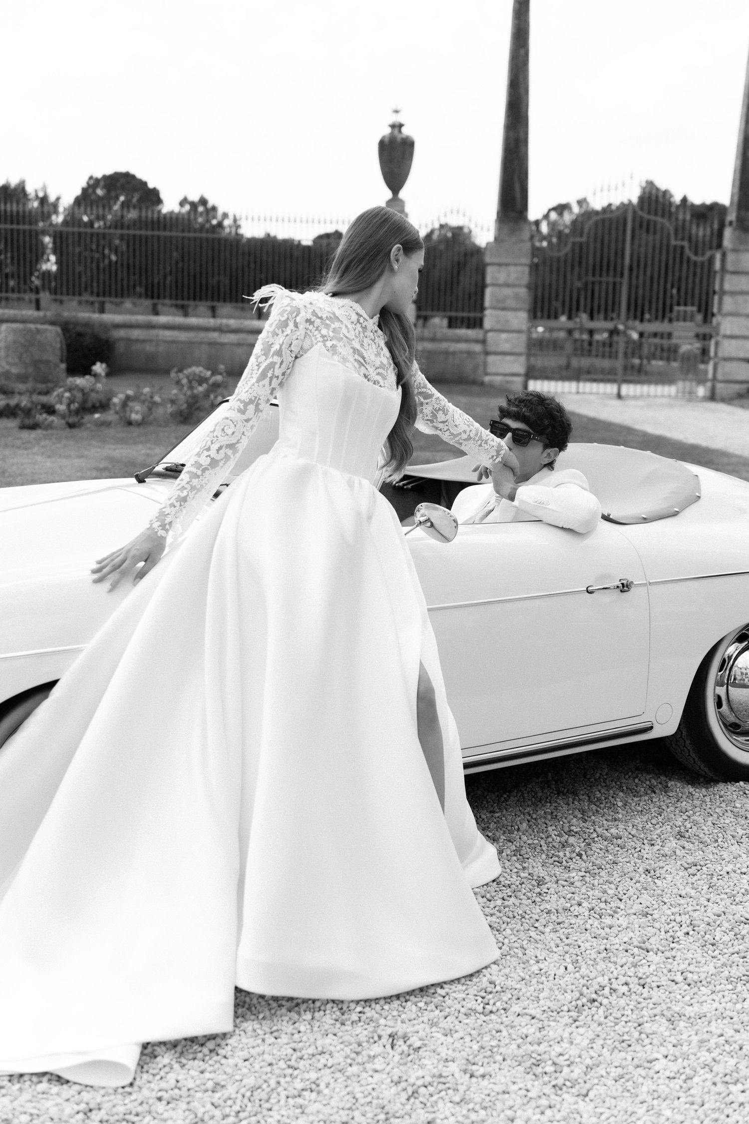 A bride in a flowing white gown poses dramatically next to a vintage white convertible car on a gravel surface. Villa Mosconi Bertani