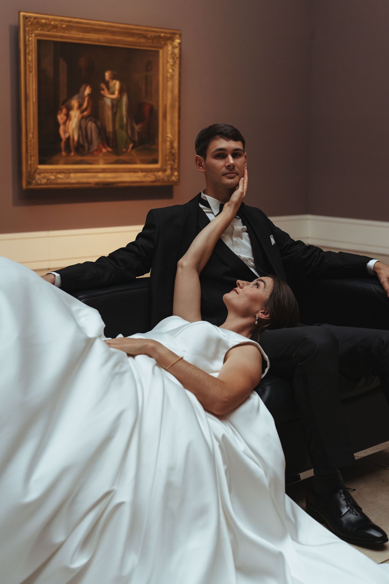 A couple in formal attire lies on the floor of an art gallery with framed paintings visible on the walls.