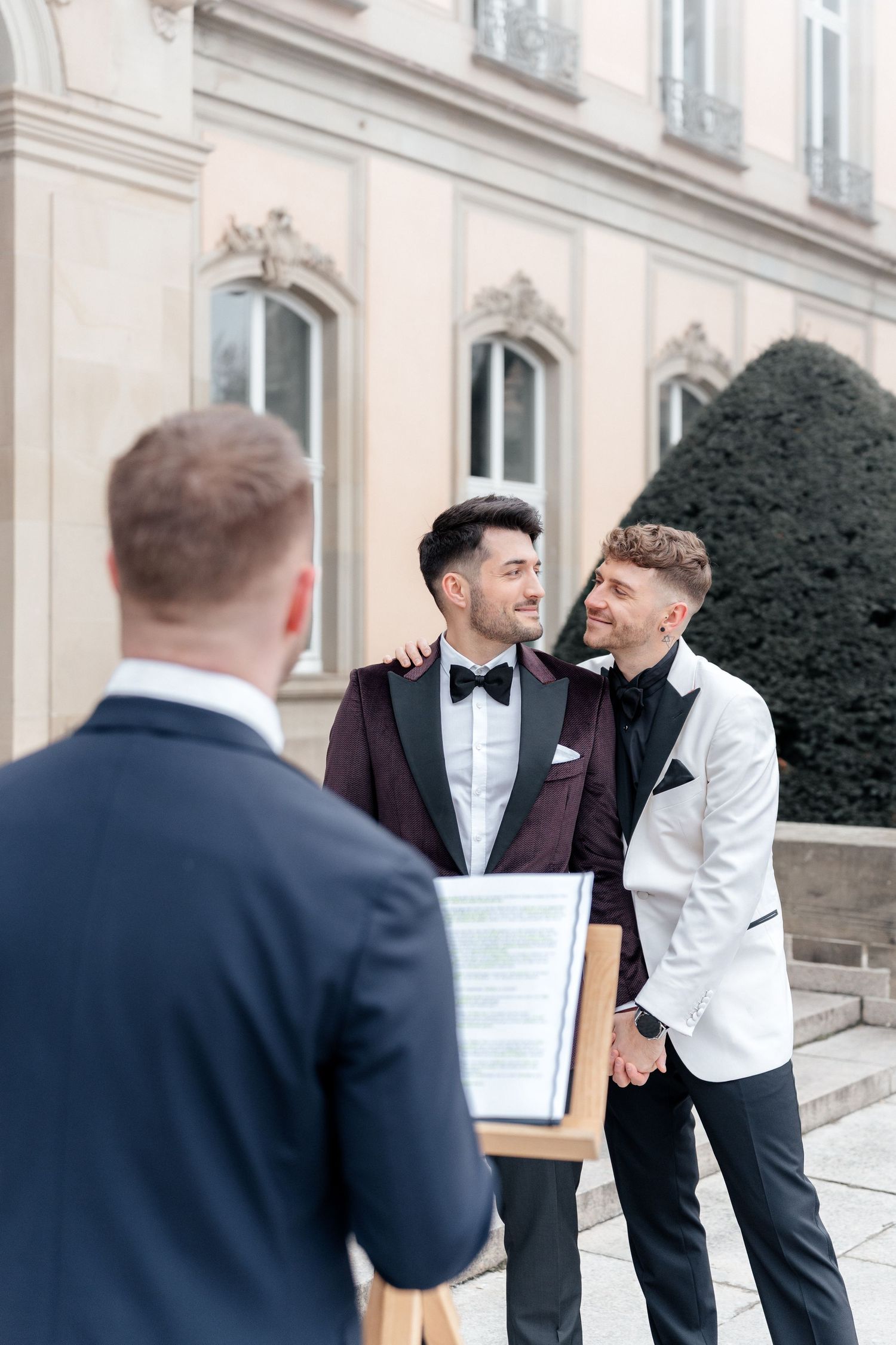 Two elegantly dressed people in tuxedos sharing a happy moment at a formal occasion while someone officiates.