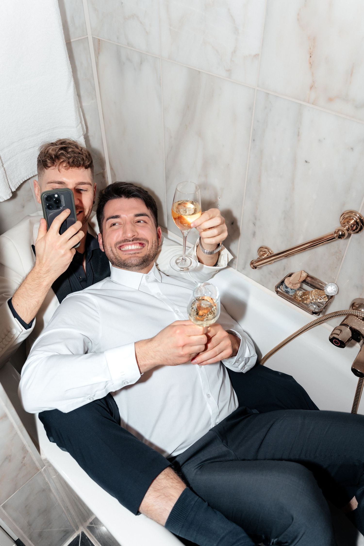 Two people relaxing in bathroom drinking wine and taking selfies with smartphones in casual attire.