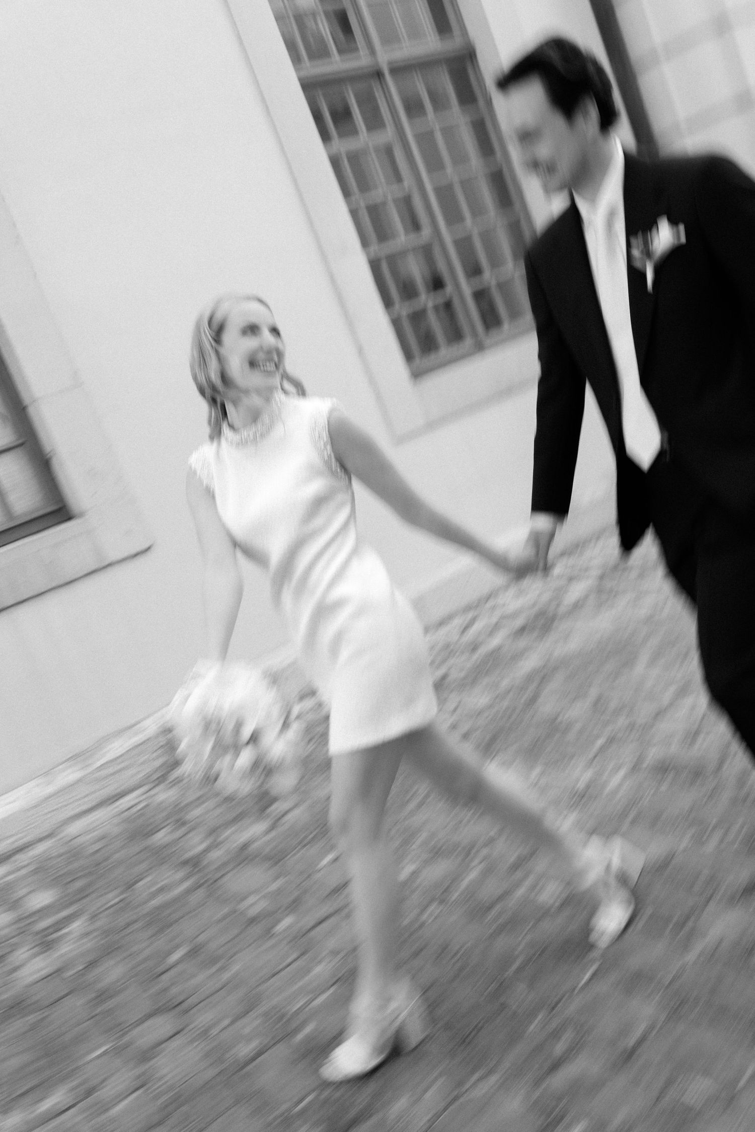 Black and white photo of a smiling bride in white dress holding hands with groom in suit, walking on cobblestone street.