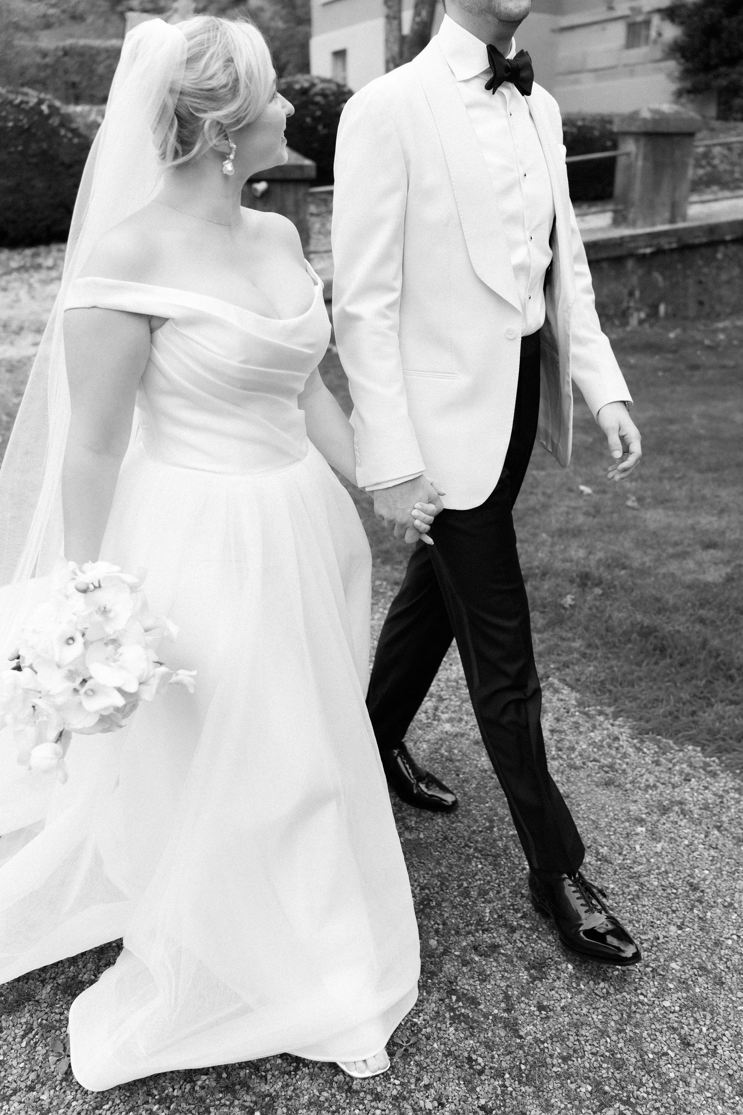 Black and white wedding photo showing a bride in a flowing gown and groom in white tuxedo jacket walking outdoors.