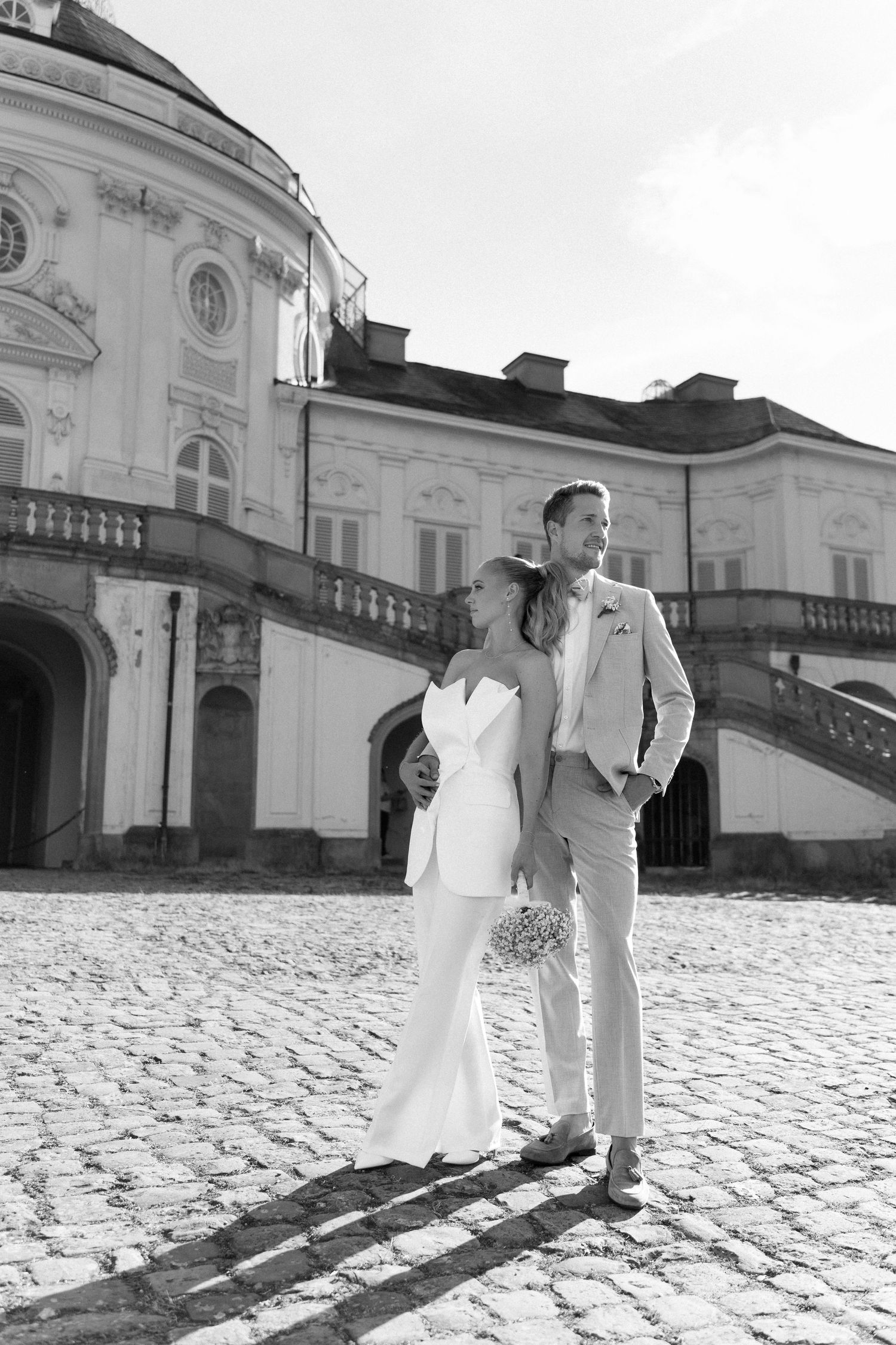 A couple in wedding attire walks along a cobblestone path beside a grand historic building with white walls. Schloss Solitude