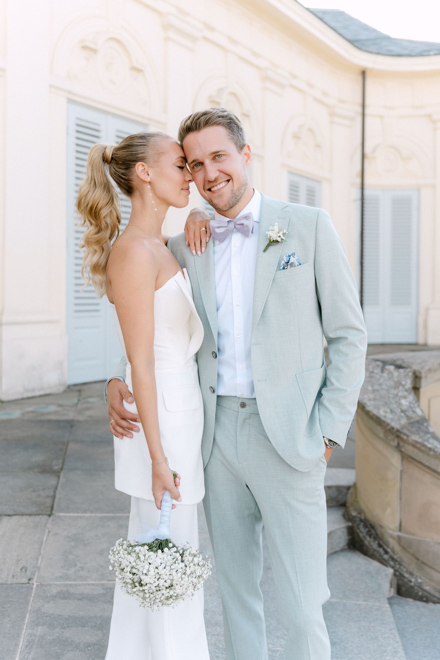 A romantic wedding couple shares intimate moments in front of a white building wearing mint green and white attire. Schloss Solitude