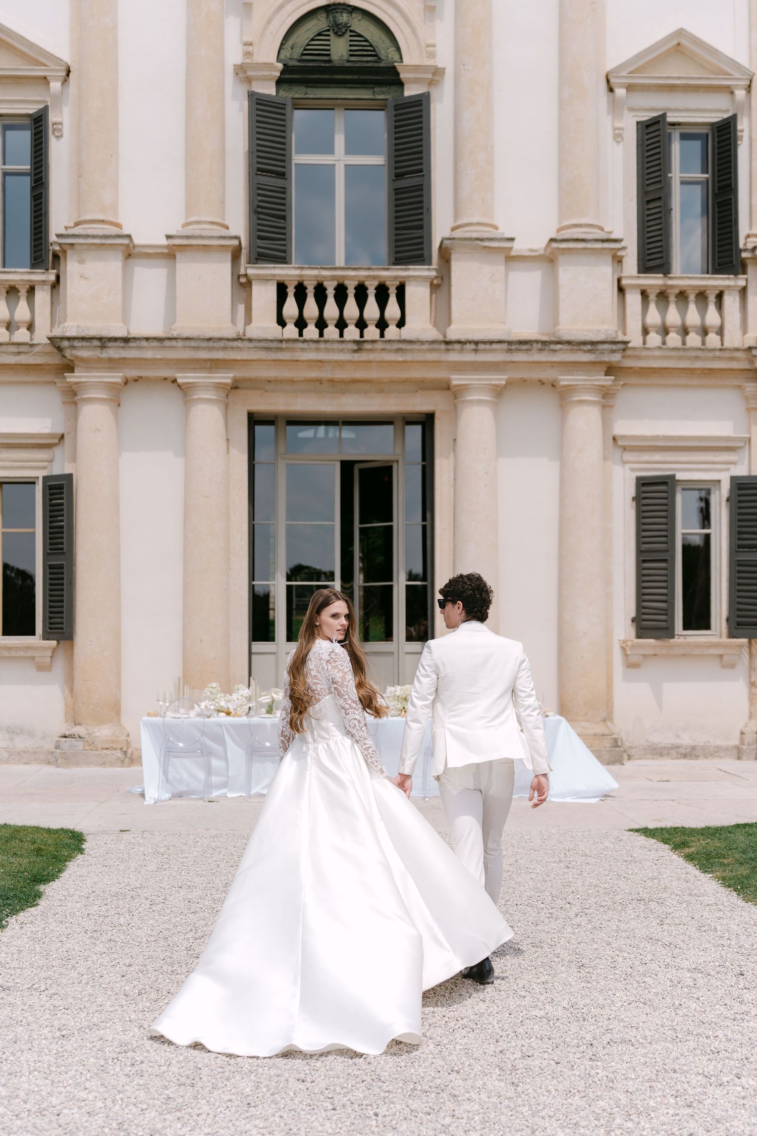Elegant couple in white attire dances gracefully across a gravel courtyard in front of a historic italian villa. Villa Mosconi Bertani