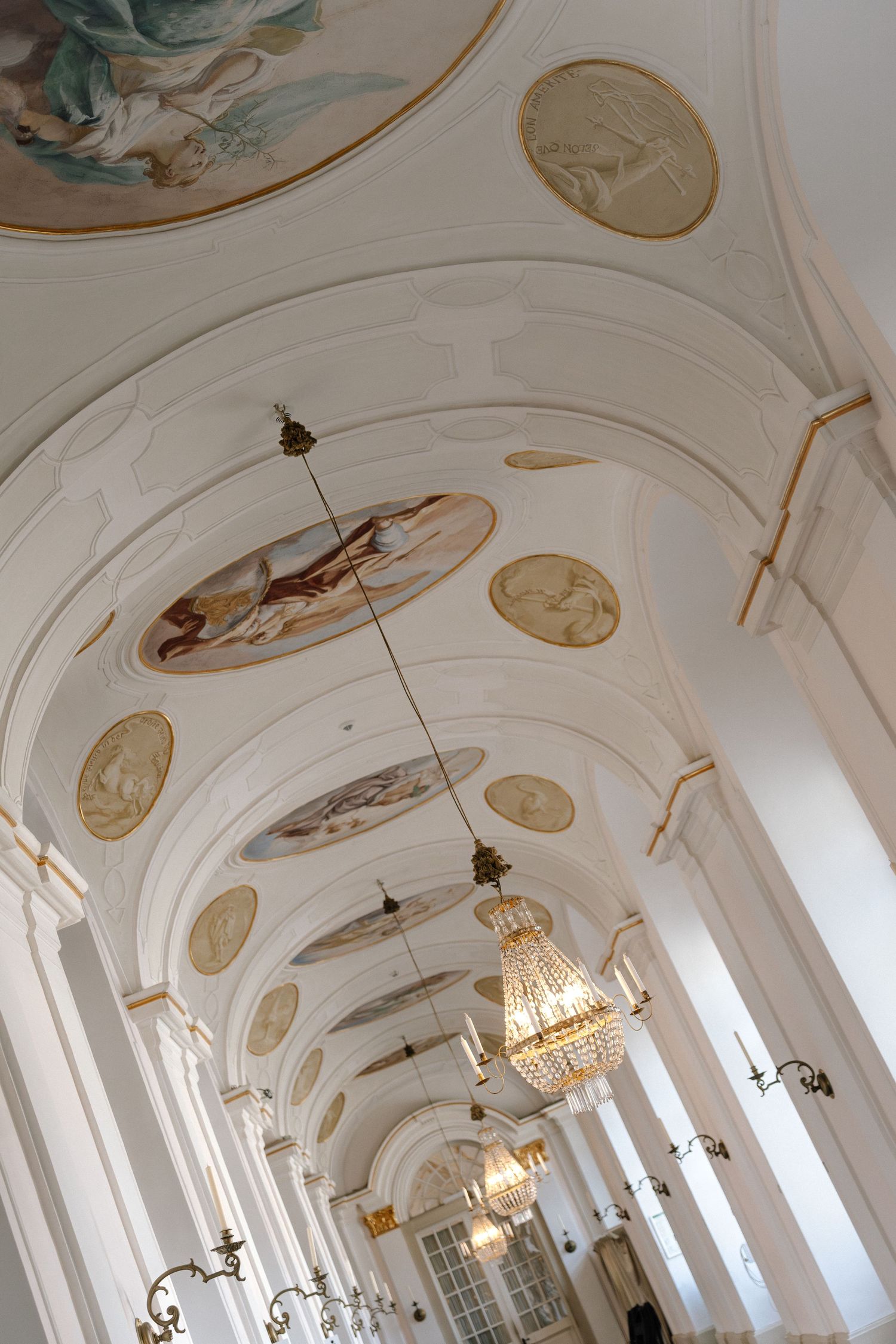 Elegant white palace corridor with ornate vaulted ceiling, crystal chandeliers, and classical architectural details. Schloss Ludwigsburg