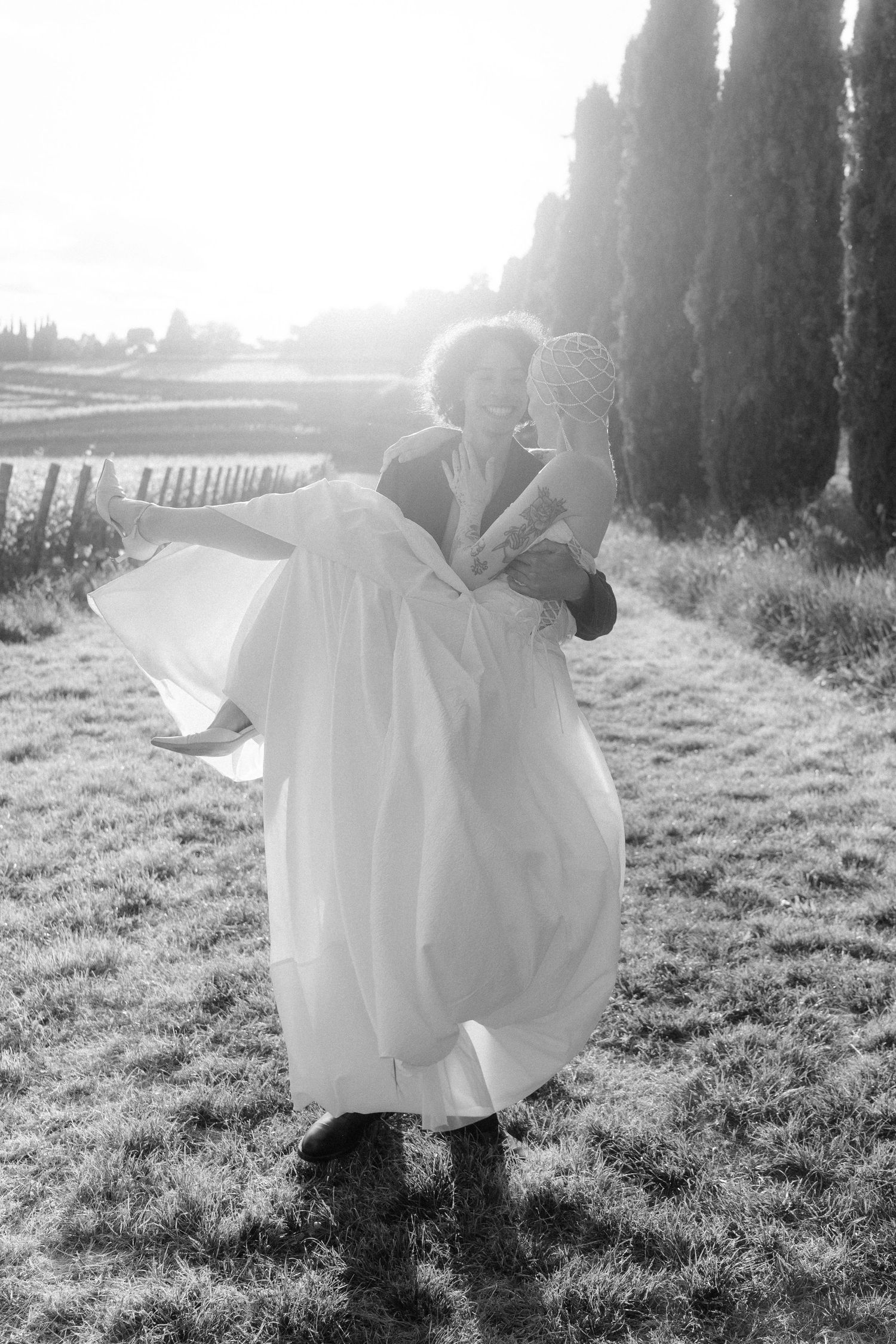 A romantic black and white wedding photo of a couple embracing in a sunlit field with cypress trees in the background.