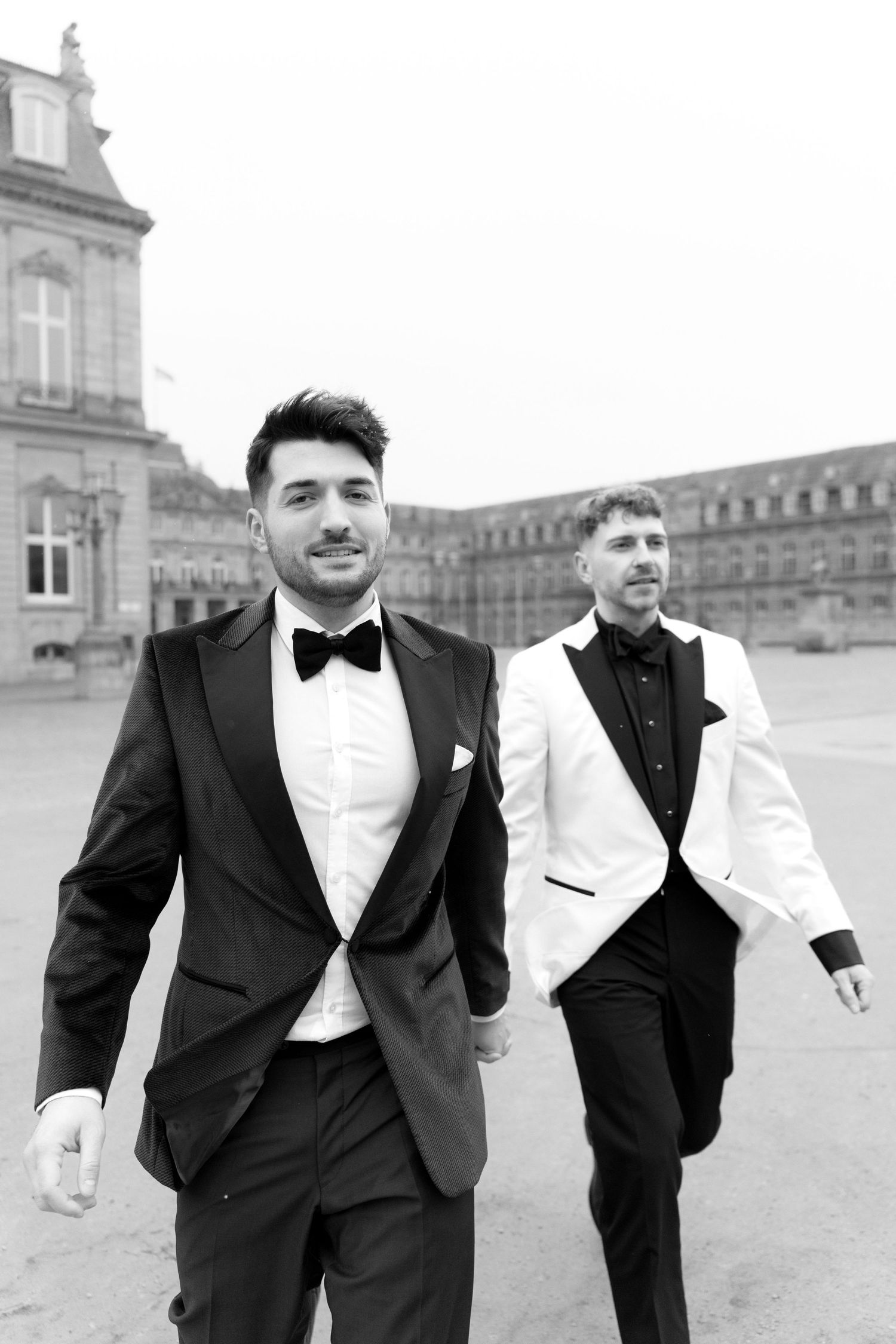 Two elegantly dressed men in formal tuxedos and bow ties walking outdoors in front of a grand classical building.