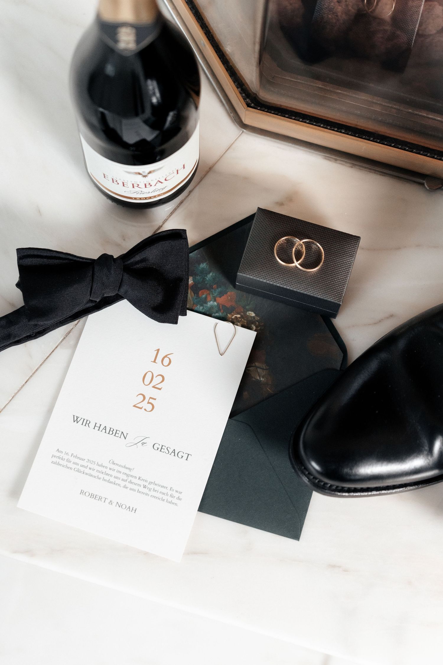 Wedding details including a black bow tie, invitation with date, rings on black card, dress shoe and champagne bottle on white surface.