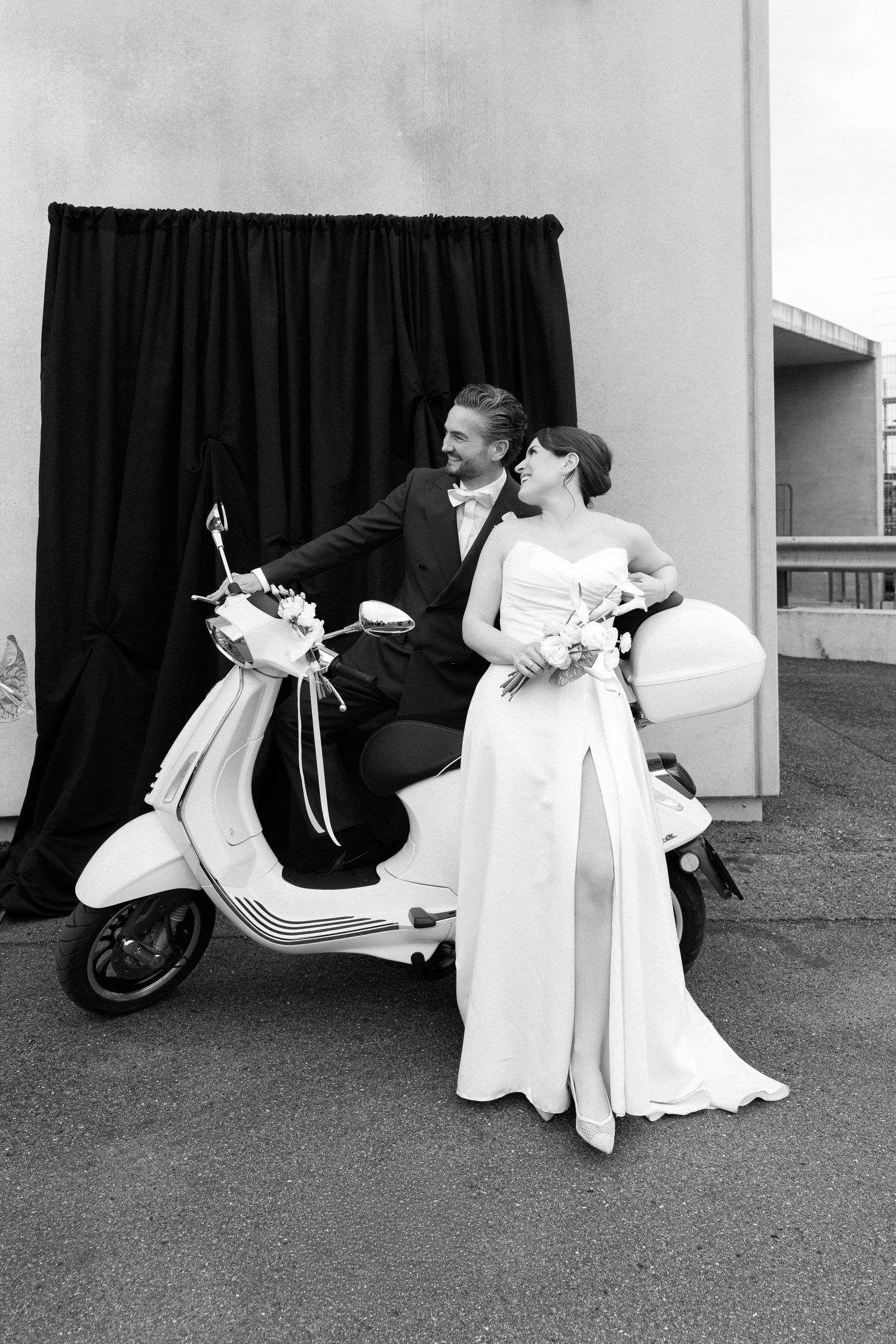Black and white photo of a bride and groom posing together on a decorated scooter in front of a dark curtain backdrop.