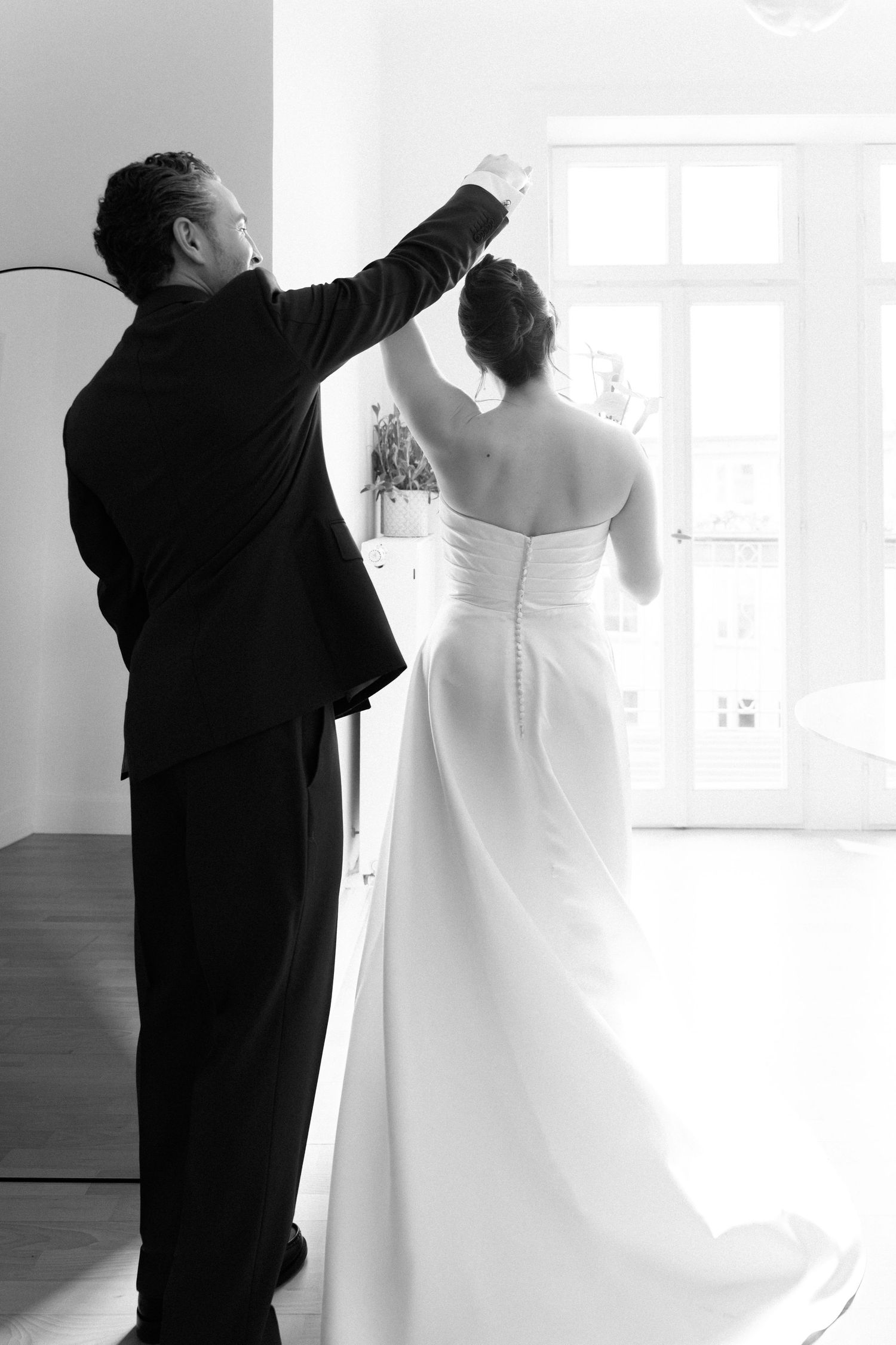 Black and white photo of a groom helping bride with her strapless wedding gown near bright window.