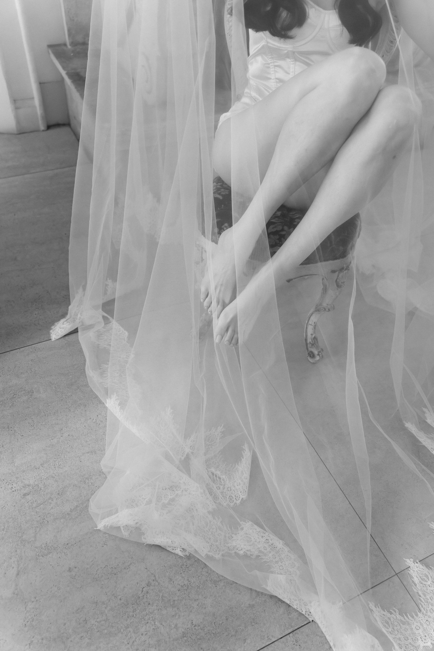 Black and white photo of a bride in lace wedding dress and veil, sitting and adjusting her bridal heels.