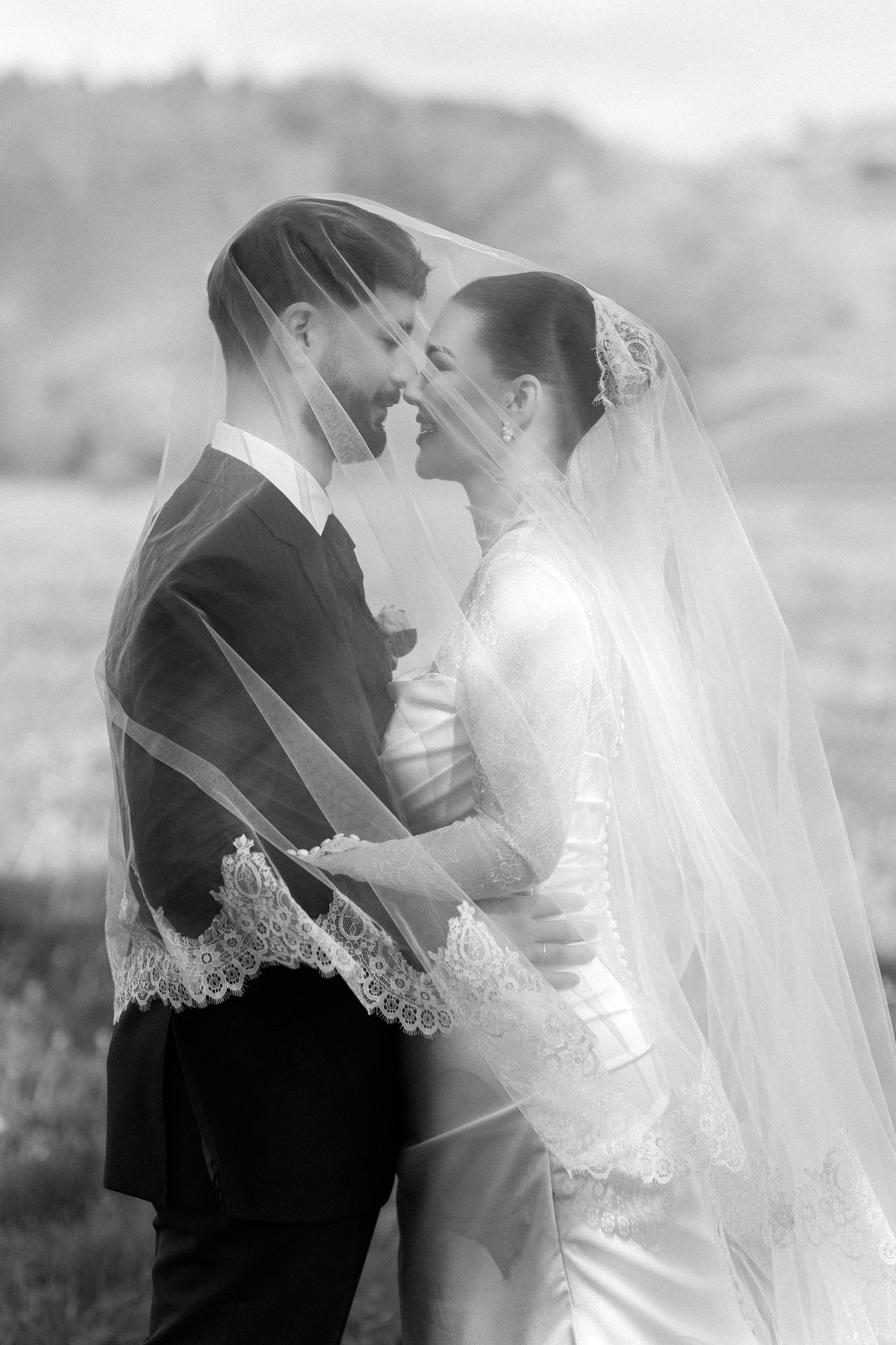 Black and white photo of bride and groom sharing intimate moment under lace wedding veil outdoors.