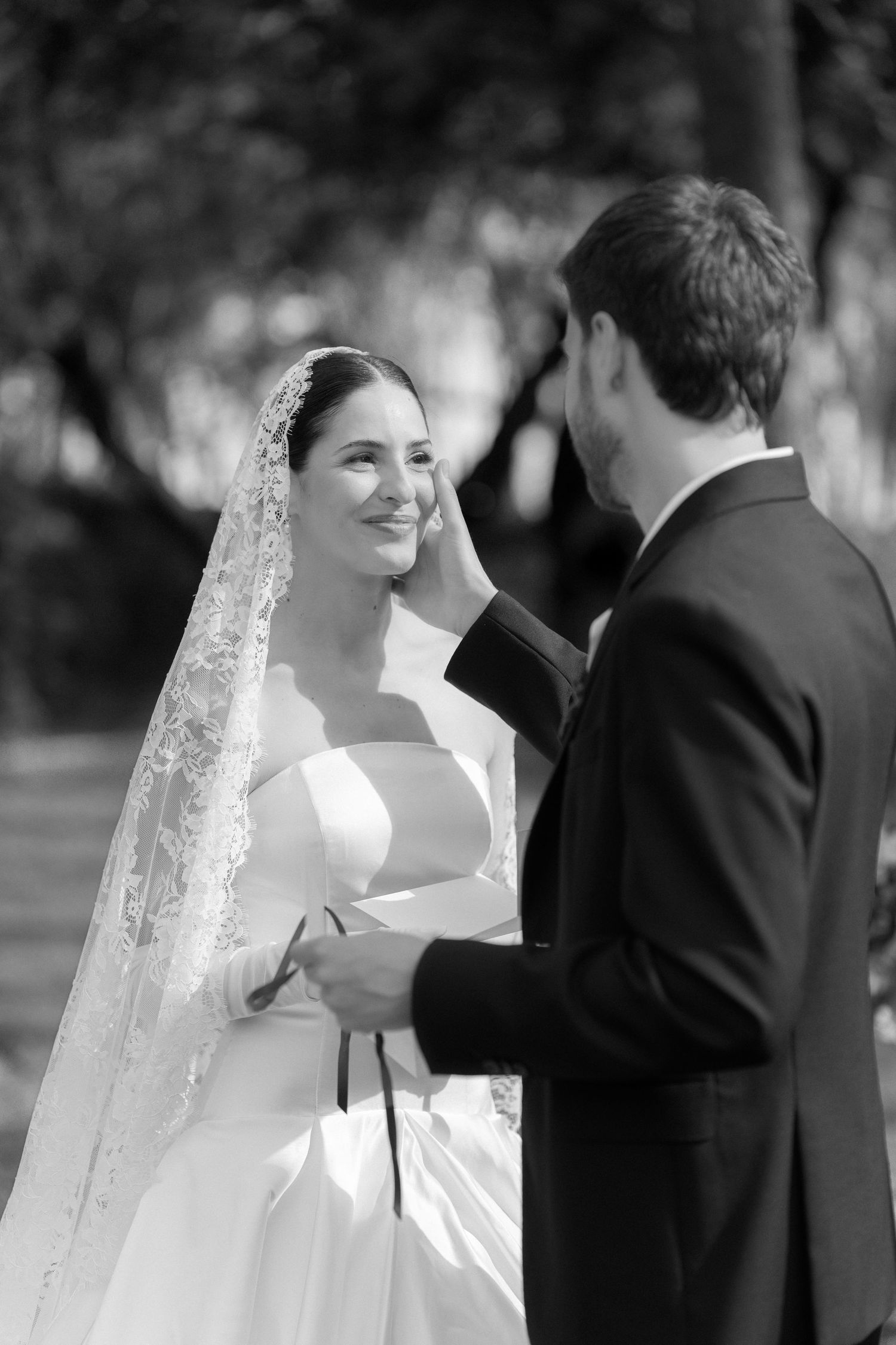 Black and white photo of a groom gently wiping a tear from his smiling bride's cheek during outdoor wedding ceremony.