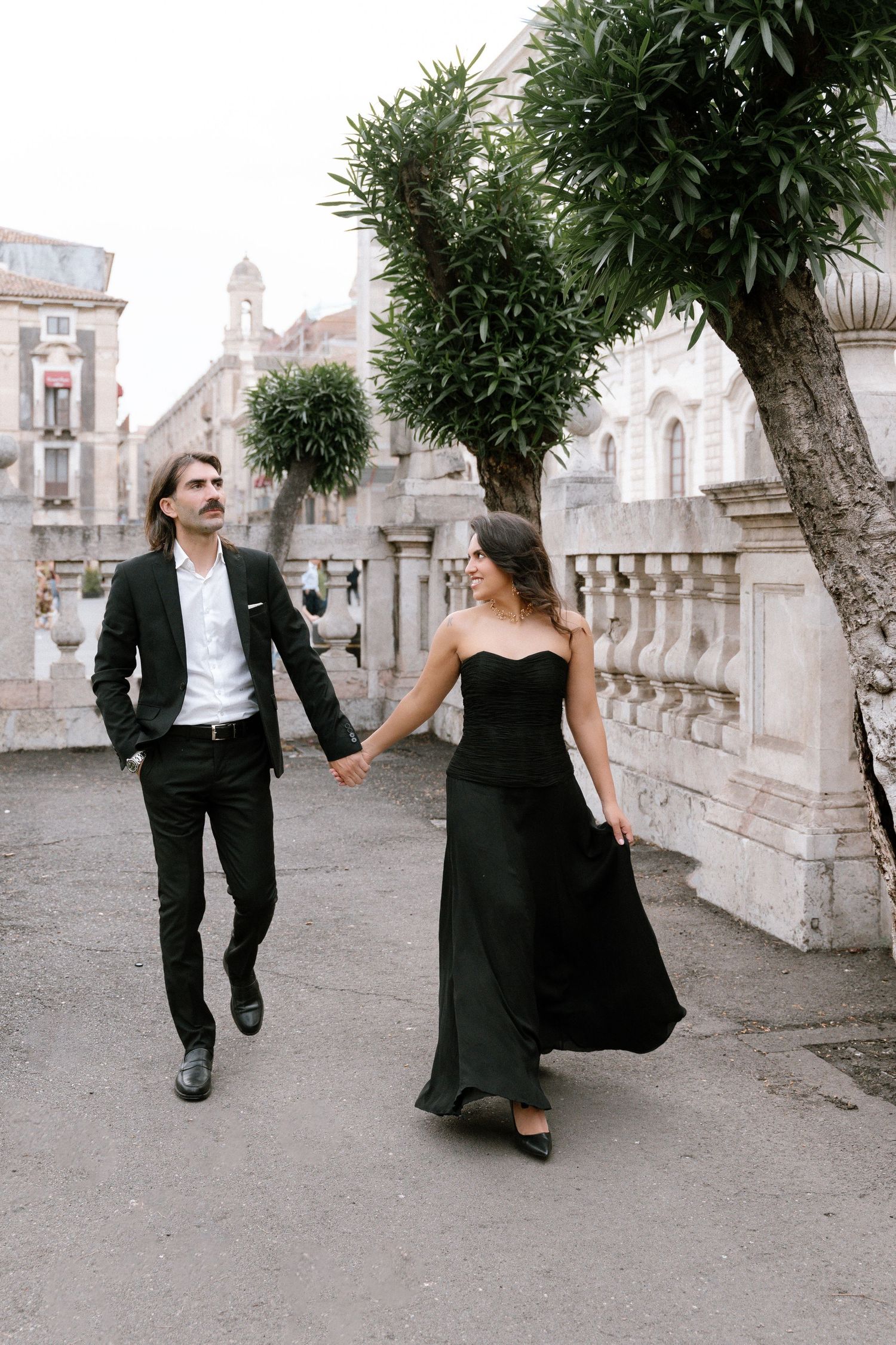 Elegant couple in matching black formal attire holding hands while walking along a classic European stone balustrade.