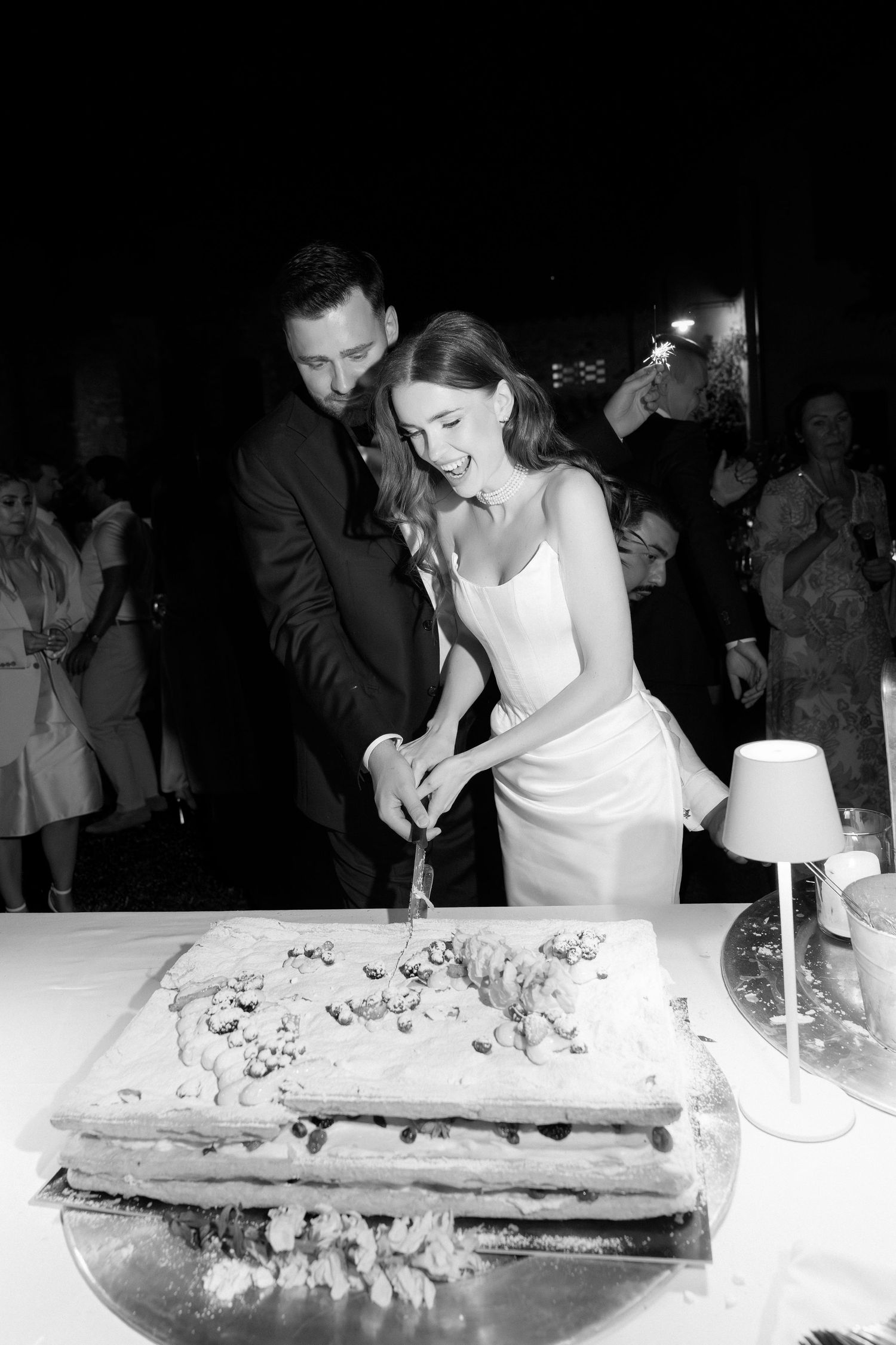 Black and white photo of a bride and groom cutting their wedding cake together at a nighttime reception.
