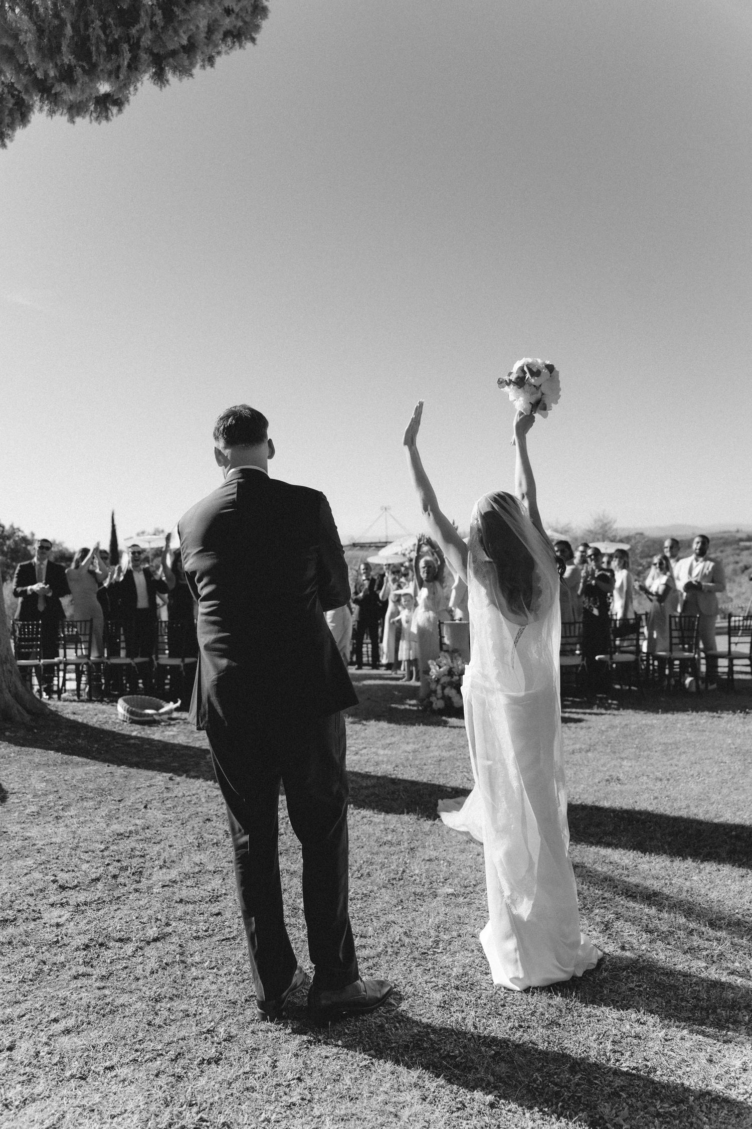 Black and white photo of bride tossing bouquet while groom stands beside her at outdoor wedding ceremony.