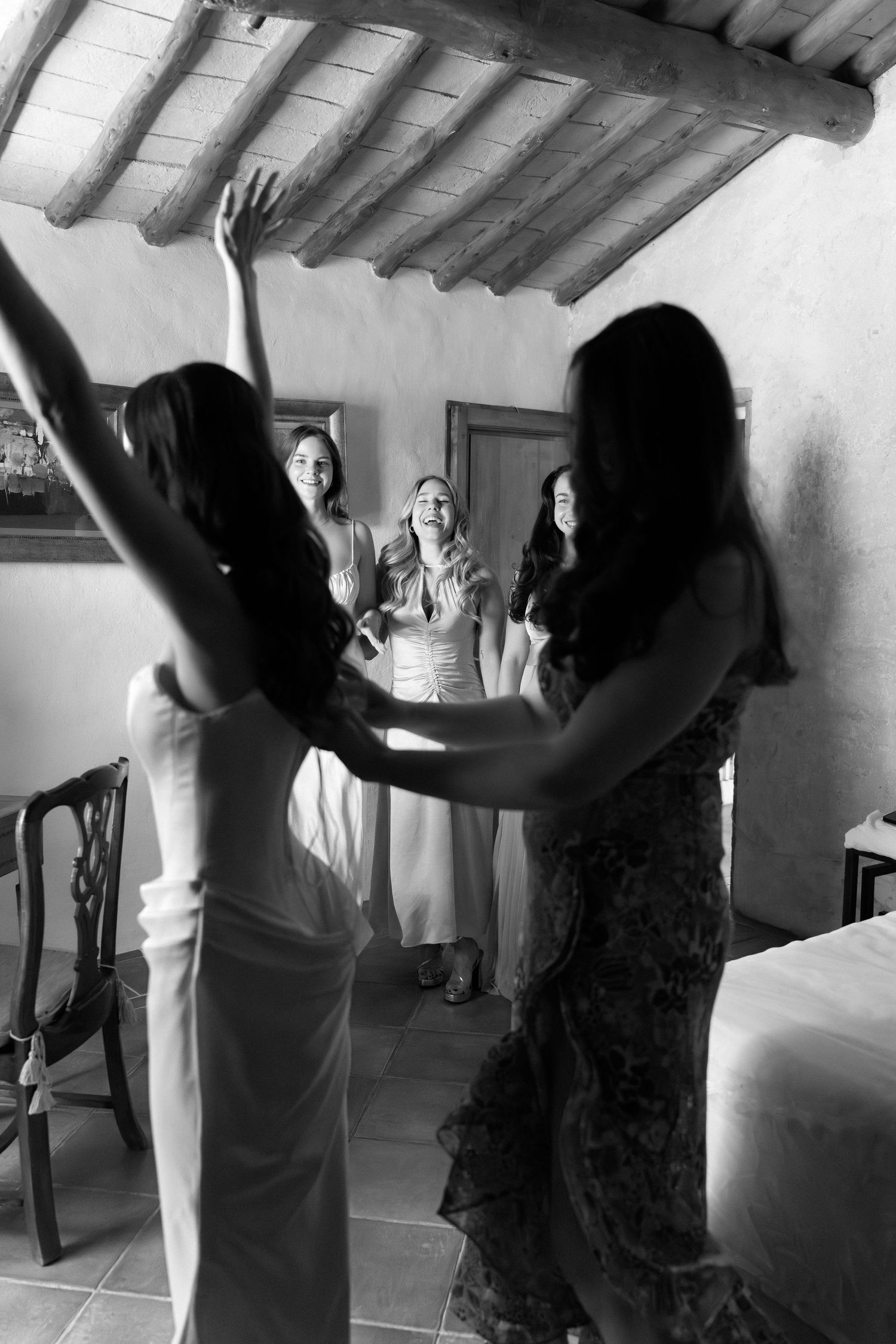 Black and white photo of bridesmaids helping bride prepare, laughing joyfully in rustic Tuscan villa room.