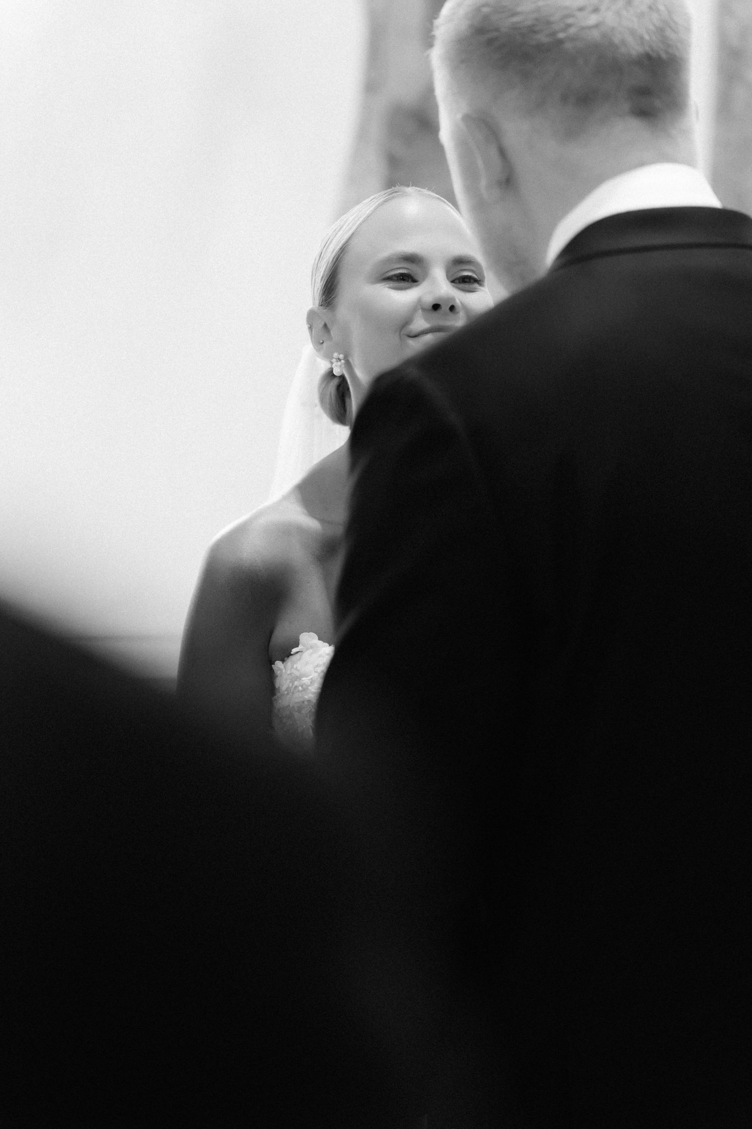 Black and white wedding photo of a smiling bride in a veil and strapless gown facing a groom in a dark suit during the ceremony.