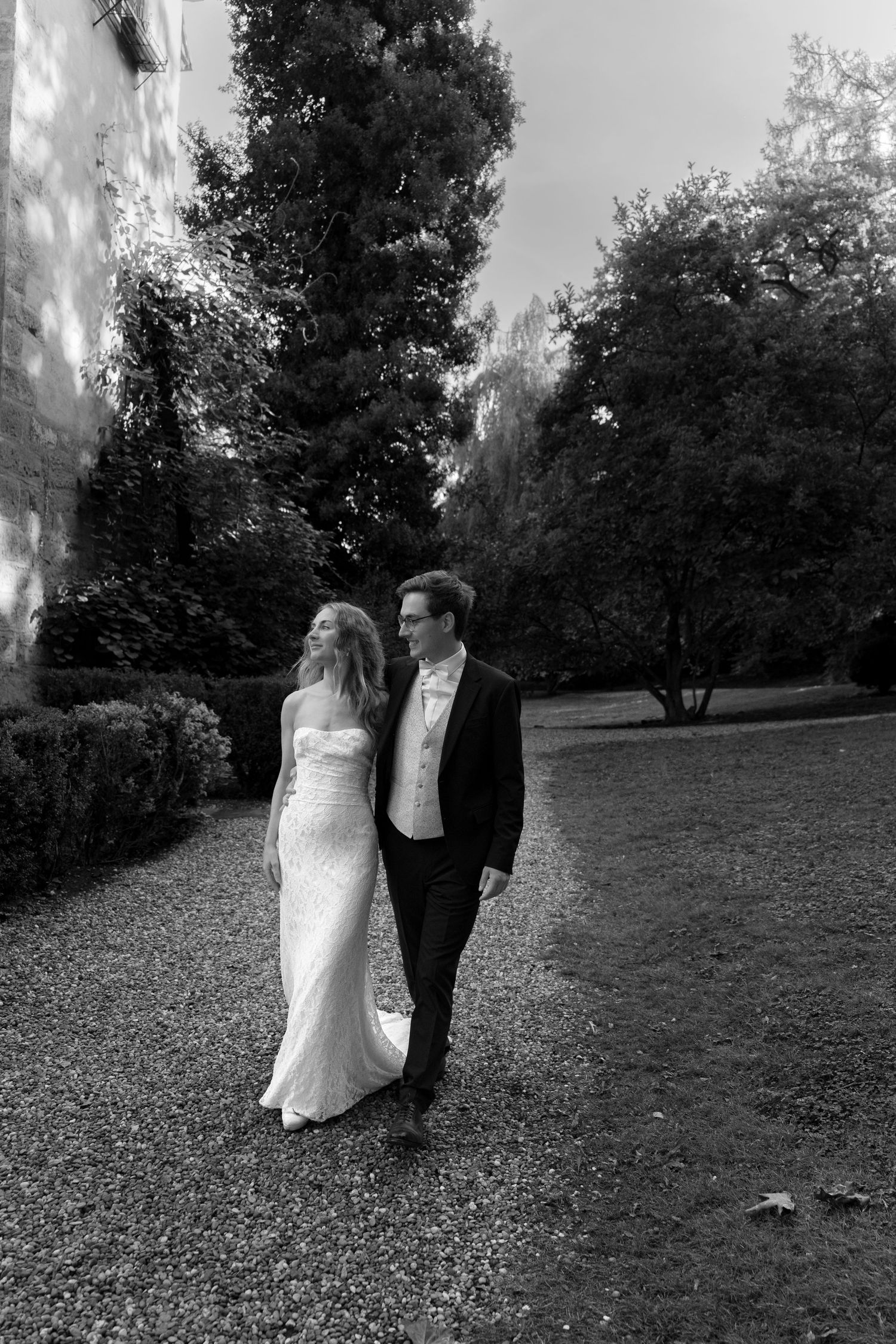 Black and white photo of a bride in a strapless lace wedding gown walking arm in arm with a groom in a suit along a tree-lined garden path.