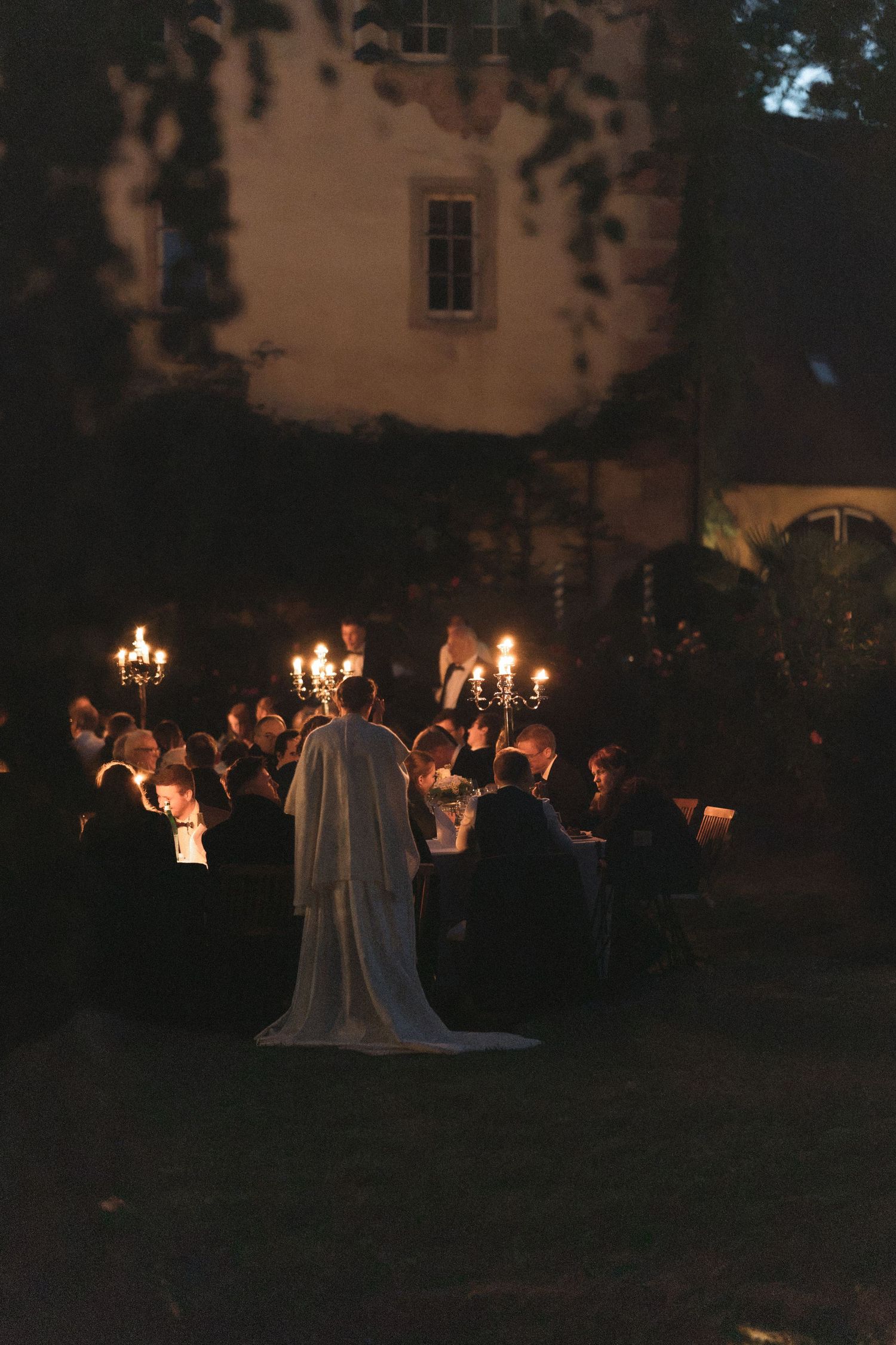 Guests gather around a candlelit dinner table in a dark garden at dusk, with a bride in a white gown standing before an old stone manor.
