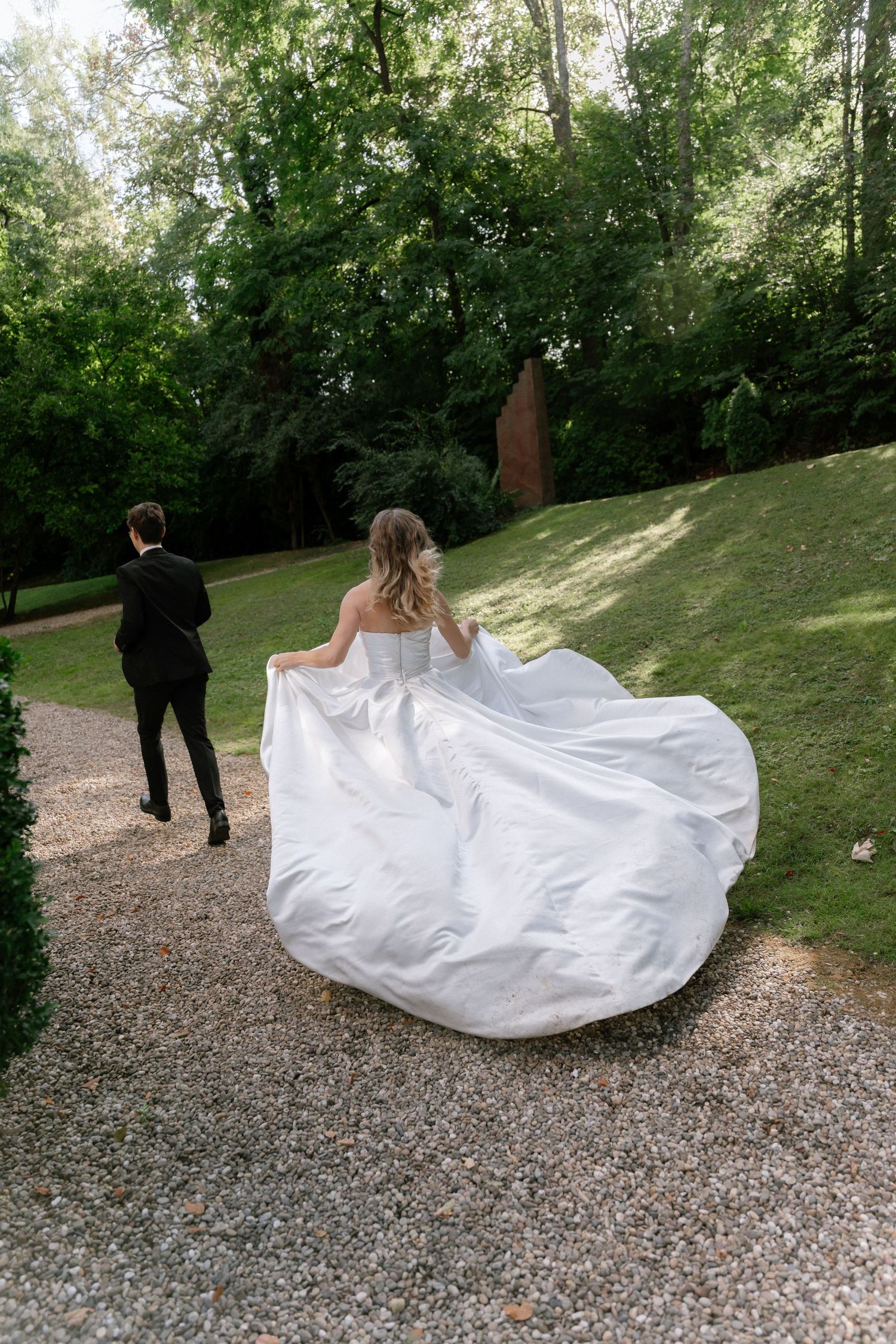 Bride in a flowing white strapless gown walks across a gravel path toward a groom in a black suit, with lush green trees and lawn behind.