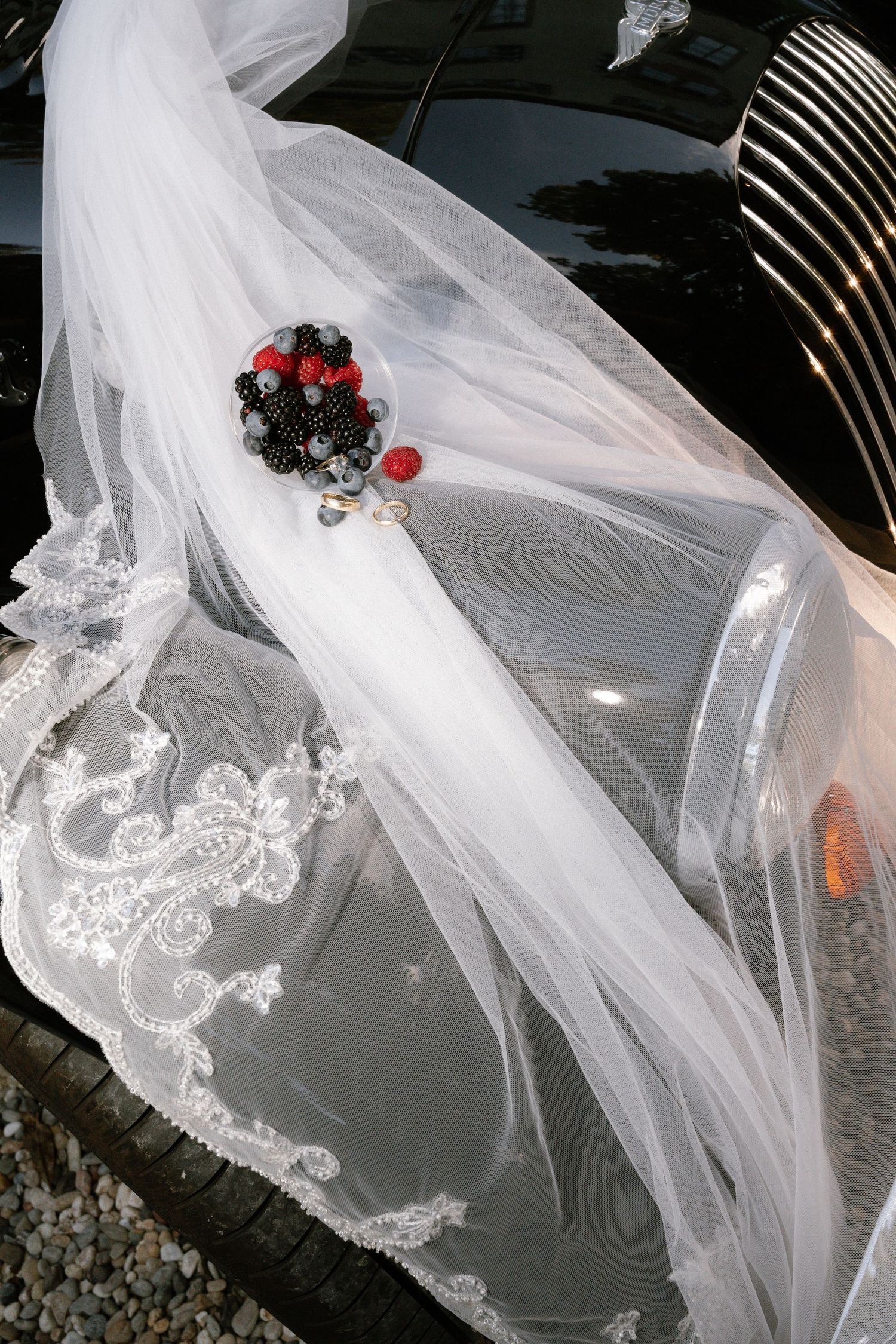 White lace-trimmed bridal veil draped over a black wedding car hood, decorated with a red and black beaded floral brooch.