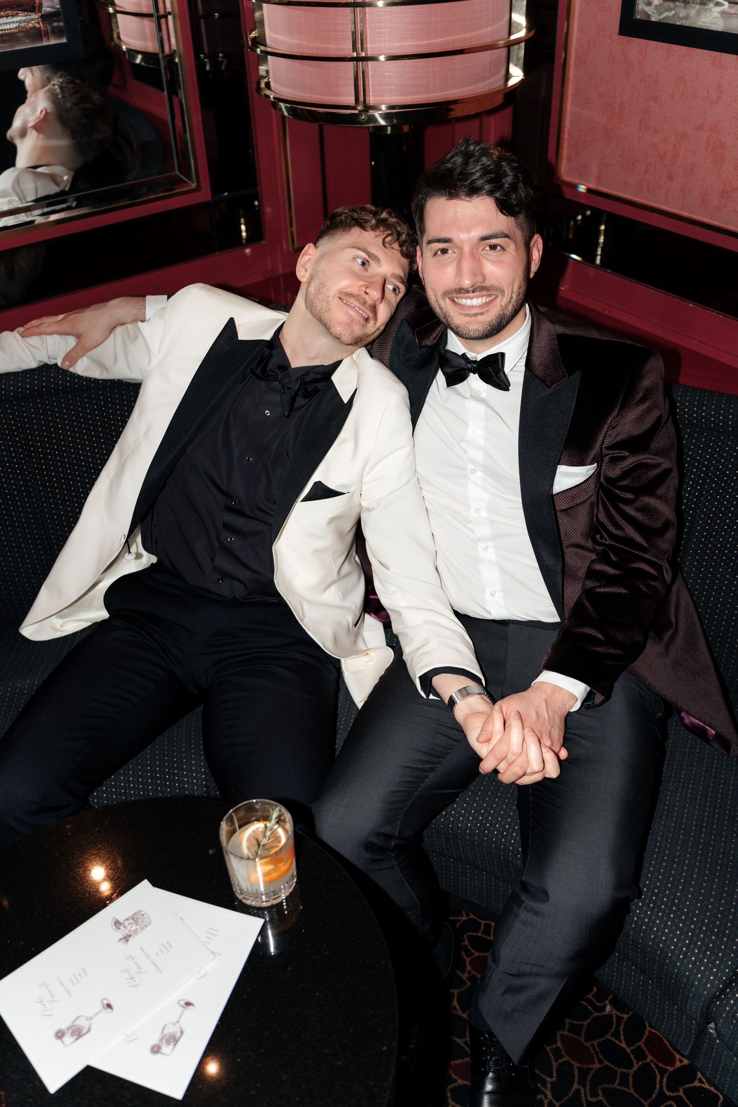 Two people in formal attire relaxing together on a red velvet banquette in a dimly lit lounge setting.