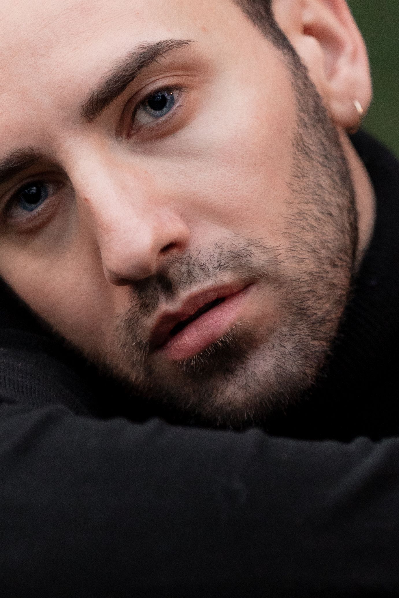 Close-up portrait photo showing intense blue eyes and facial hair against dark clothing and blurred background.