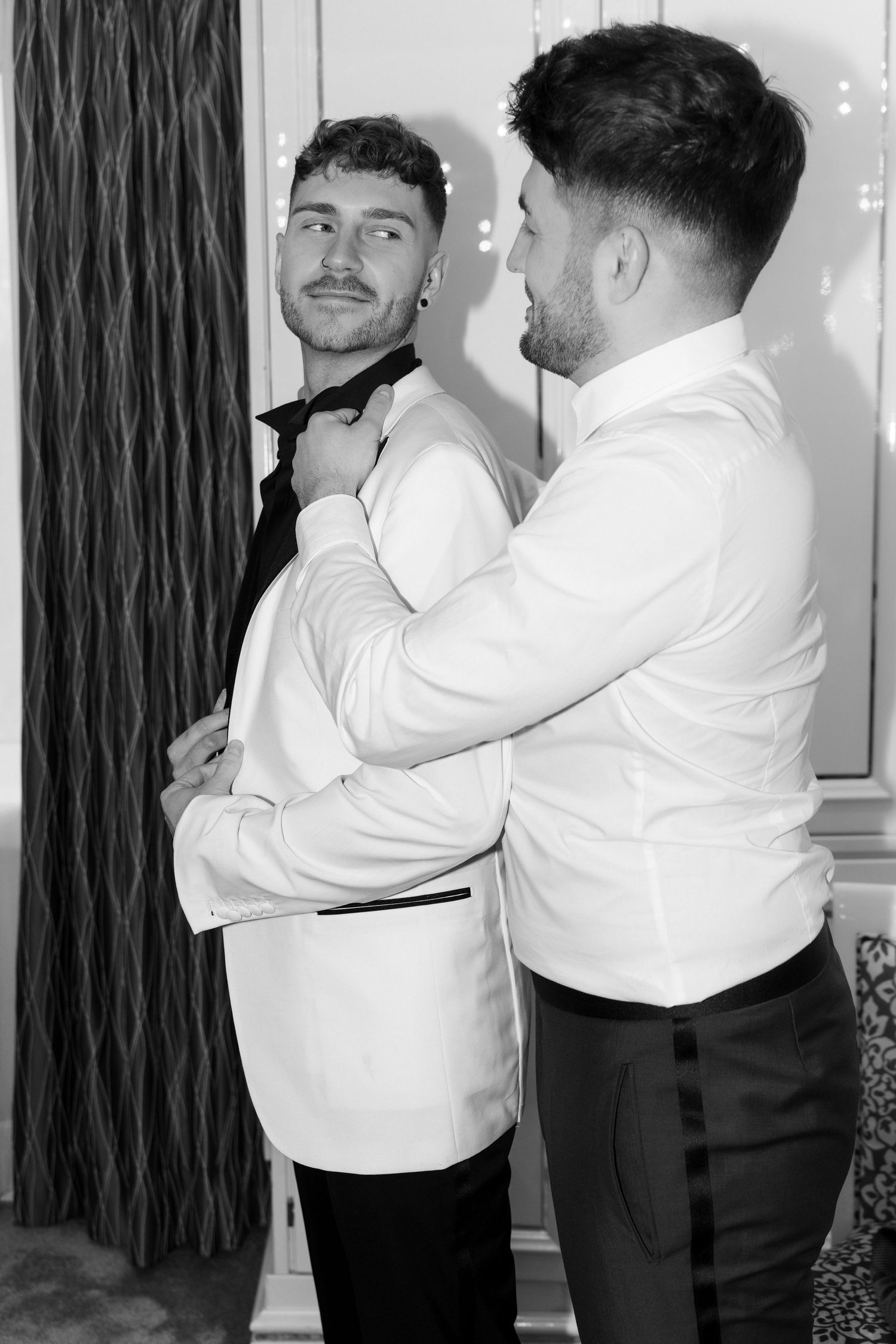 Two men in formal white dinner jackets help each other get dressed before an event in a black and white photo.
