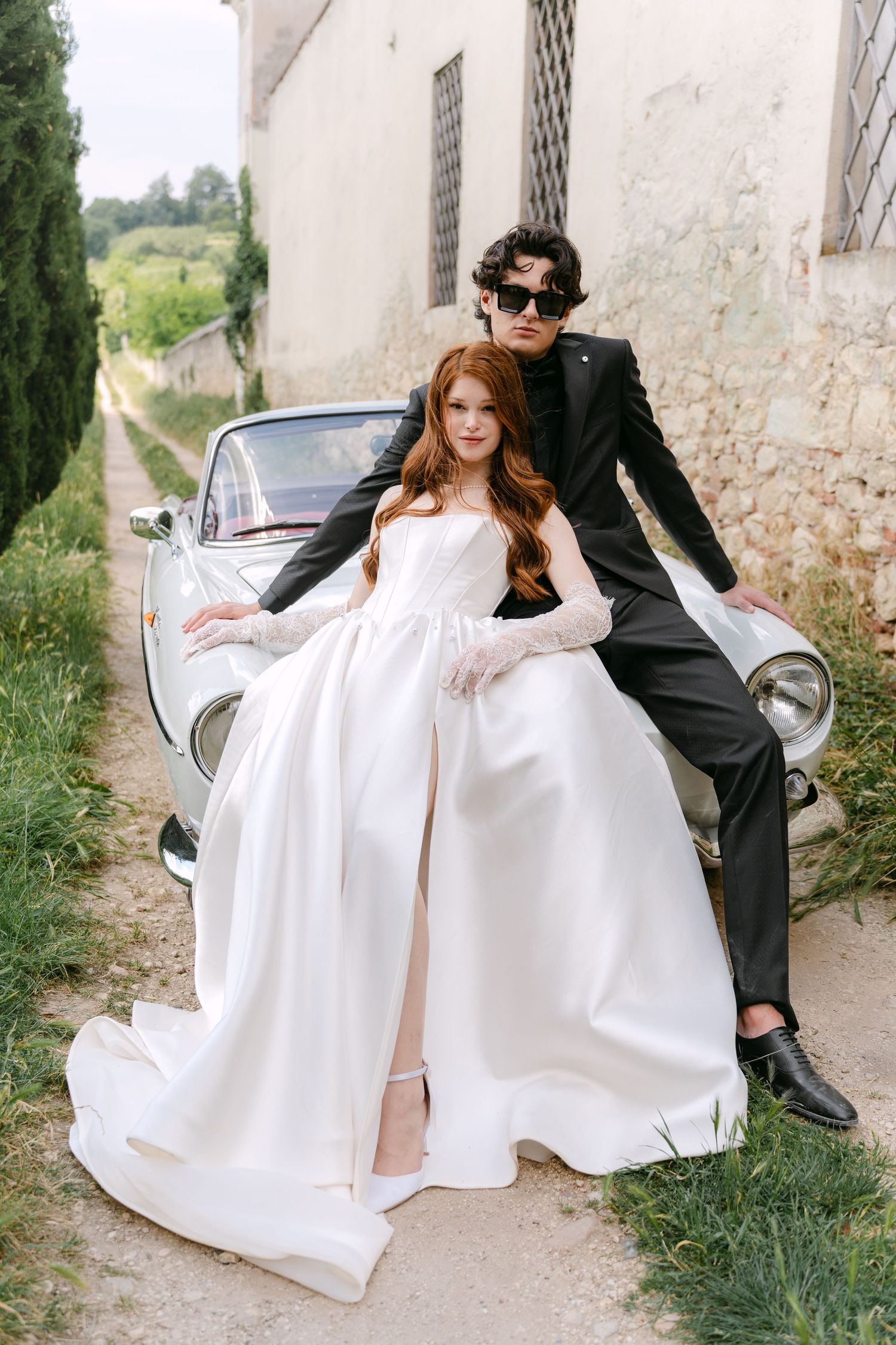 Stylish couple poses with vintage car against white stone wall in romantic wedding photo shoot.