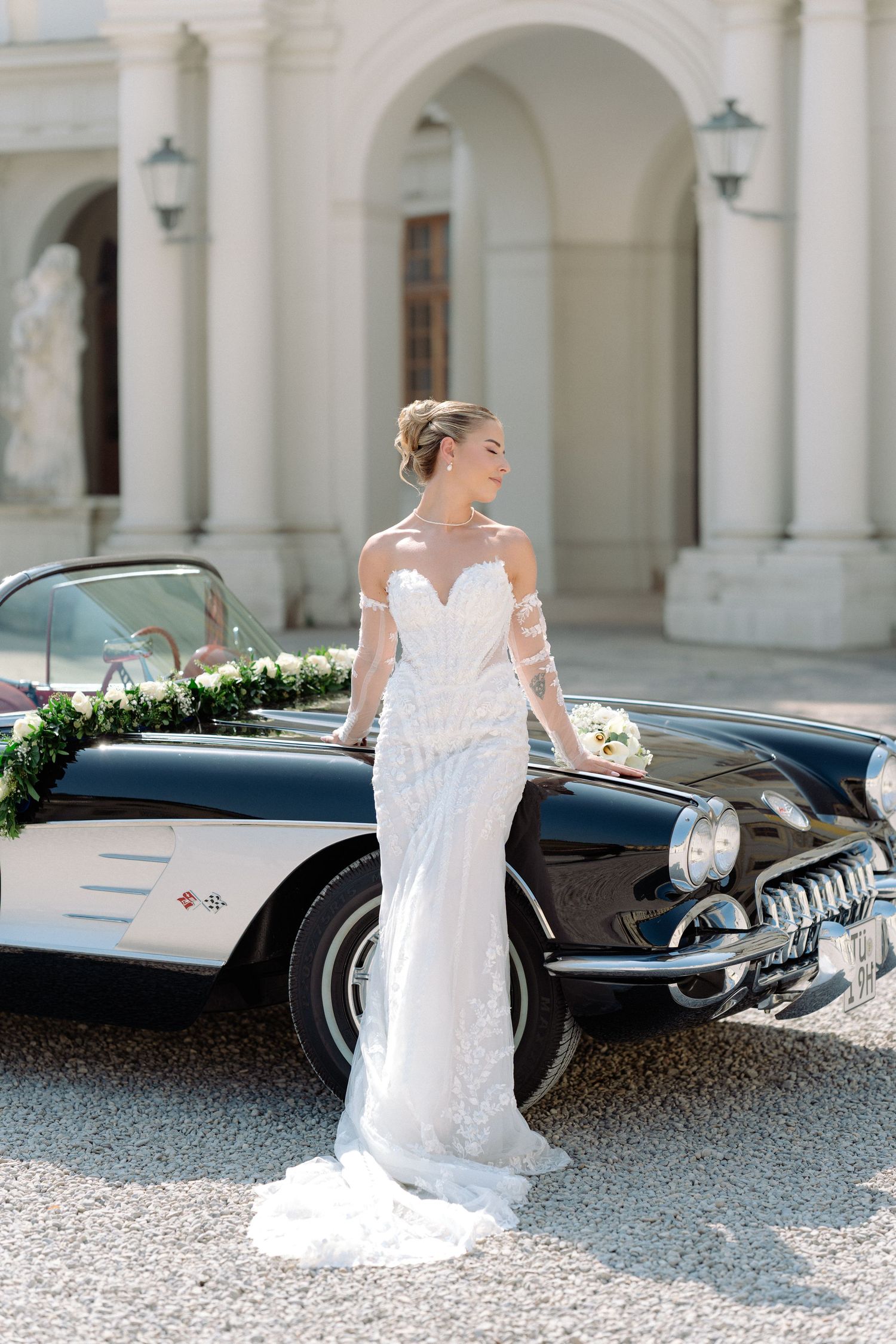 Bride in white lace dress poses elegantly next to vintage black car in front of classical arched building. Schloss Ludwigsburg