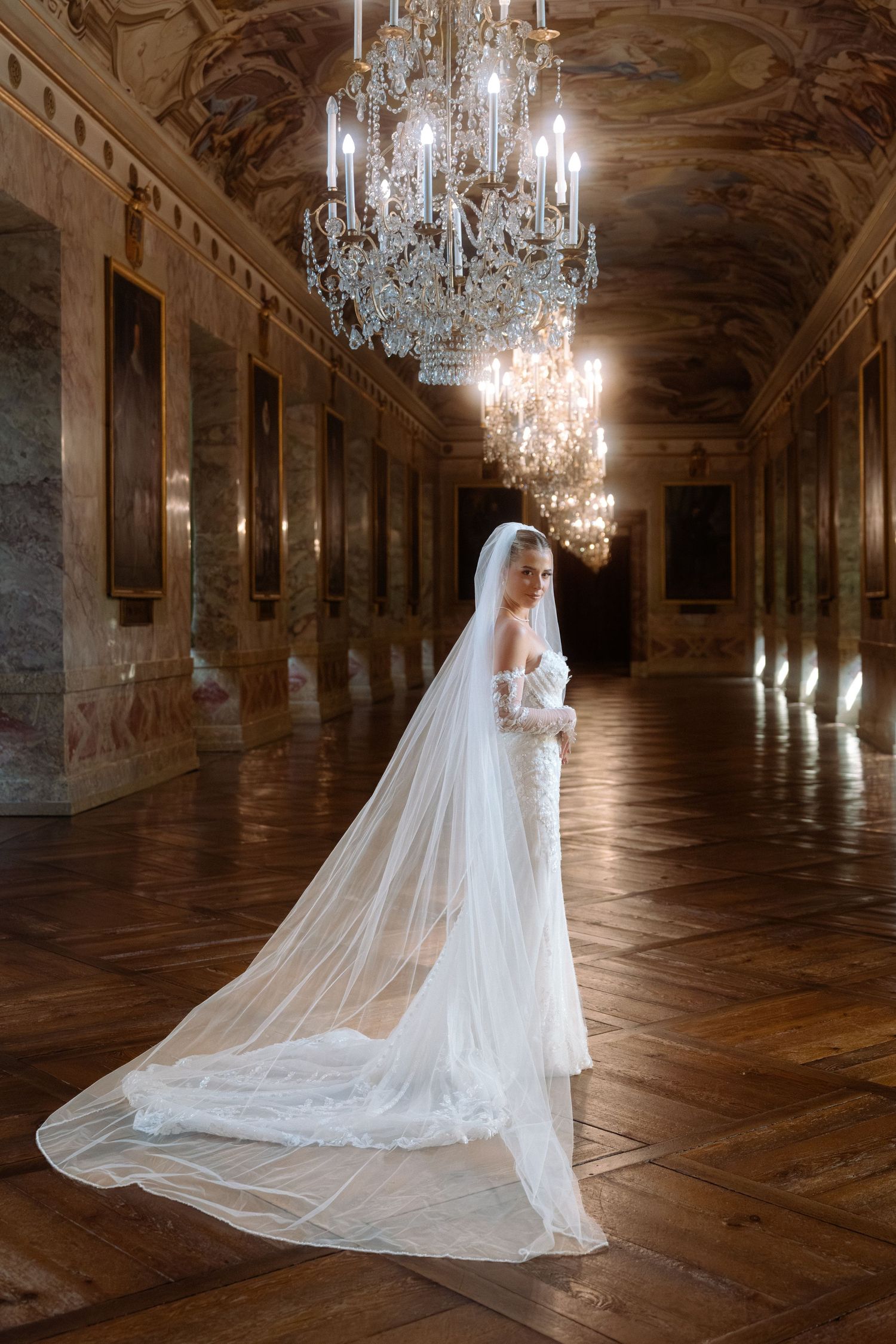 A wedding dress and long veil flow elegantly in an ornate palace hall with crystal chandeliers and polished wood floors. Schloss Ludwigsburg