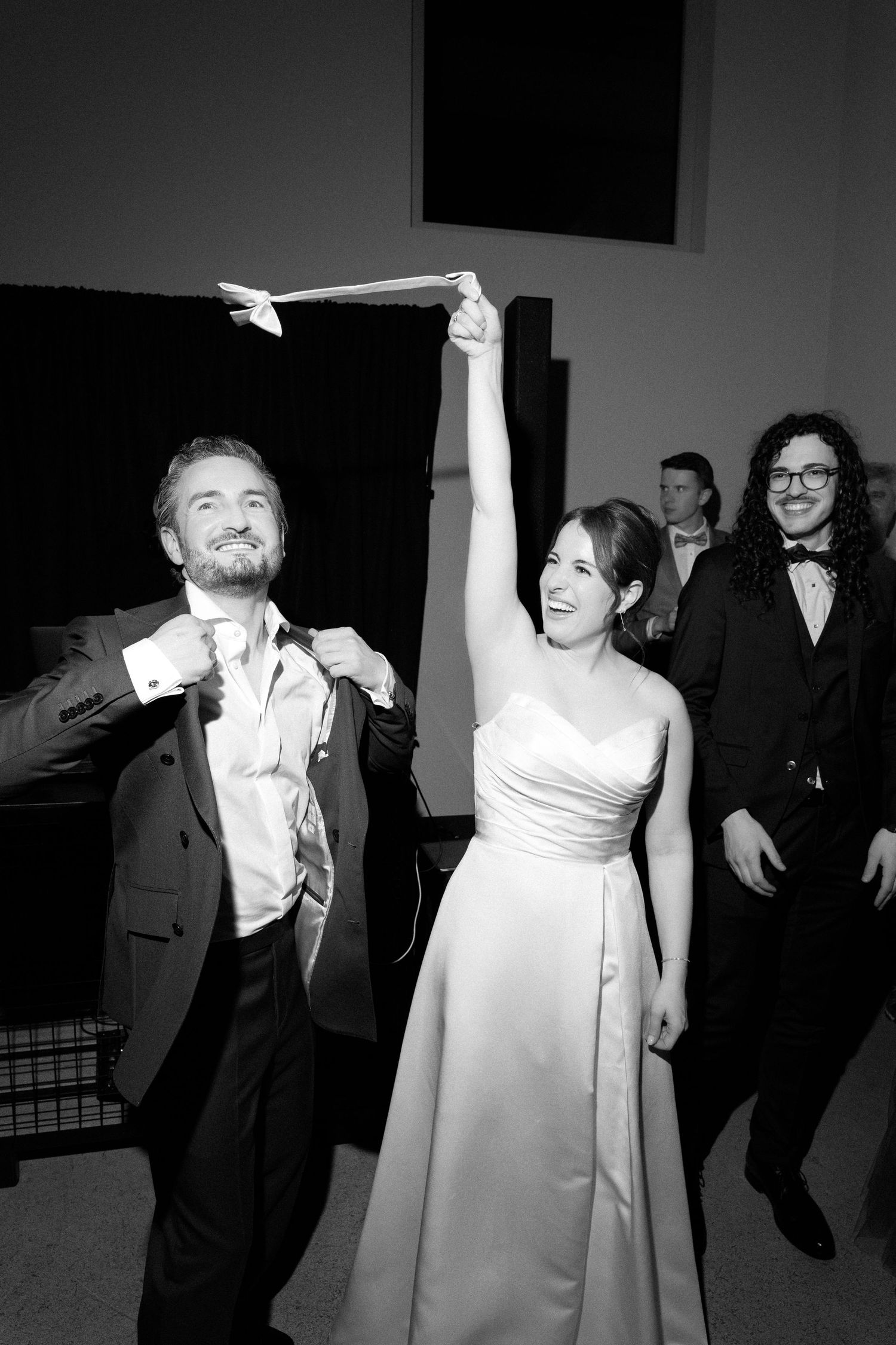 Black and white photo of a bride raising her arm joyfully beside groom at wedding reception.