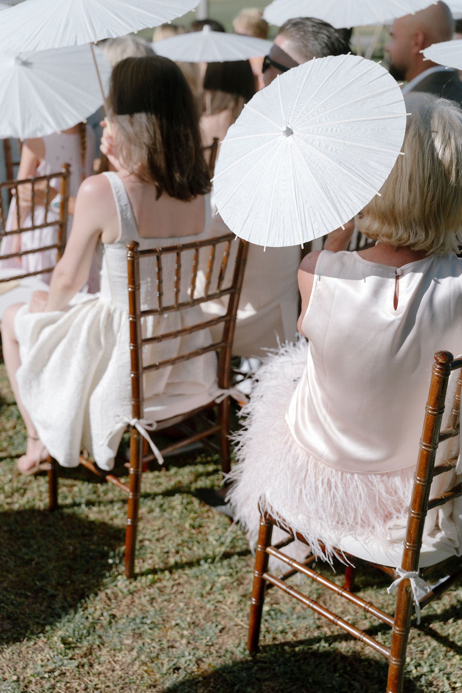 Guests seated in chiavari chairs holding white parasols at an outdoor wedding ceremony.