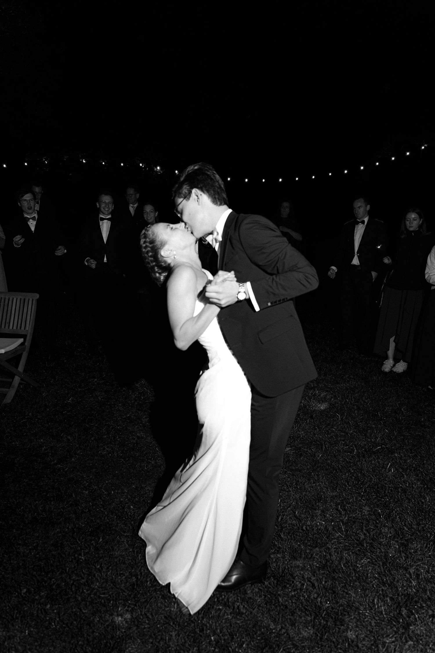 Black and white photo of a bride and groom sharing a dipped kiss during an outdoor evening wedding dance as guests watch.