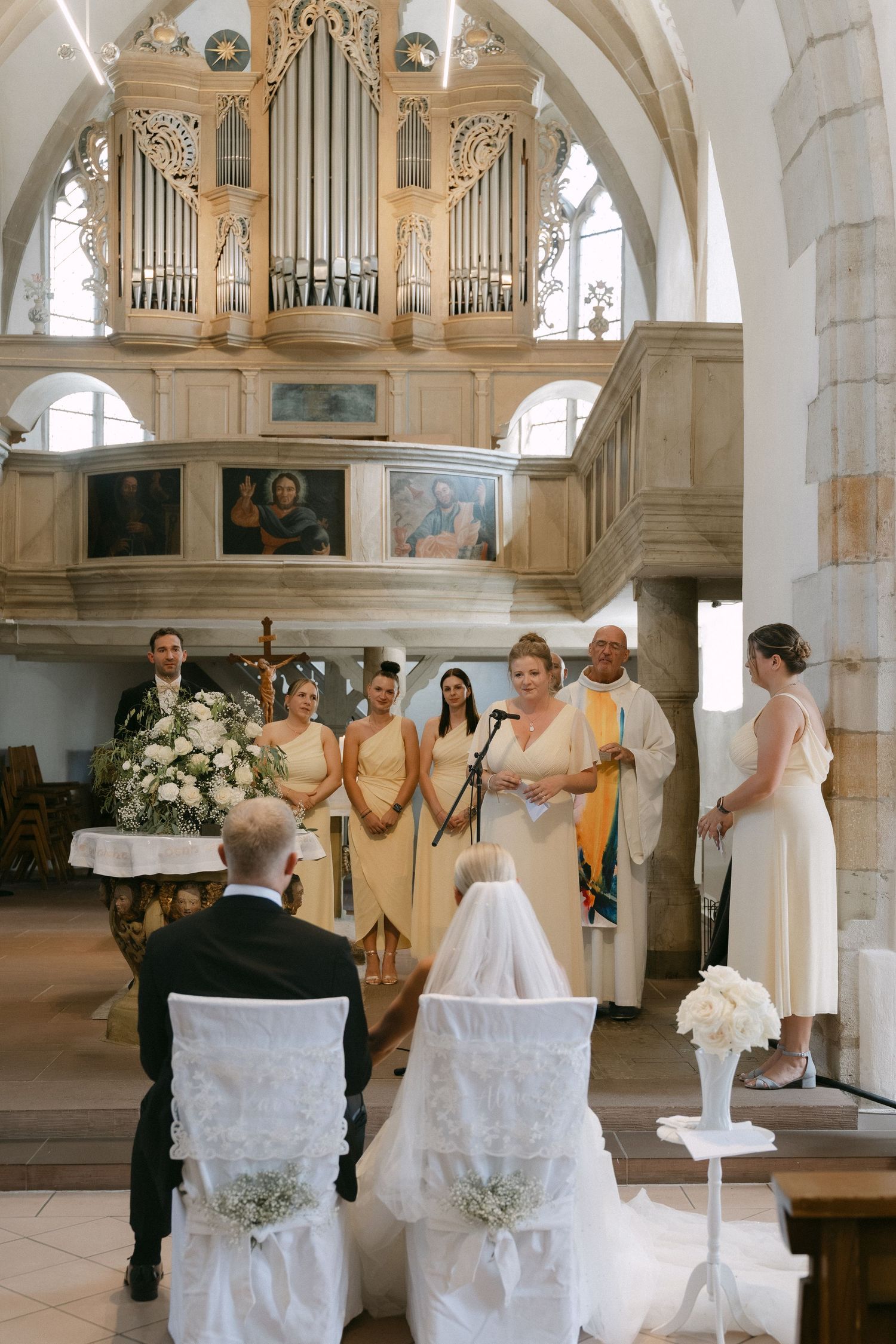 Bride and groom seated before a priest during a church wedding ceremony as bridesmaids in yellow gowns stand beneath an ornate pipe organ.
