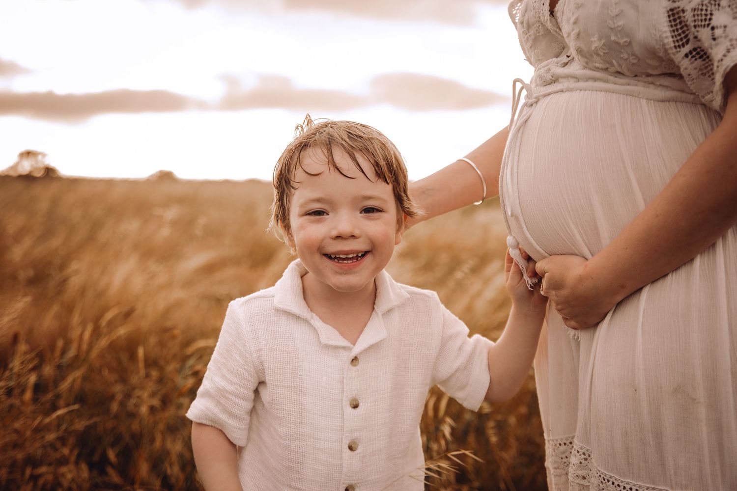 Child in white clothing running through golden field during sunset photoshoot. Pregnancy, Maternity