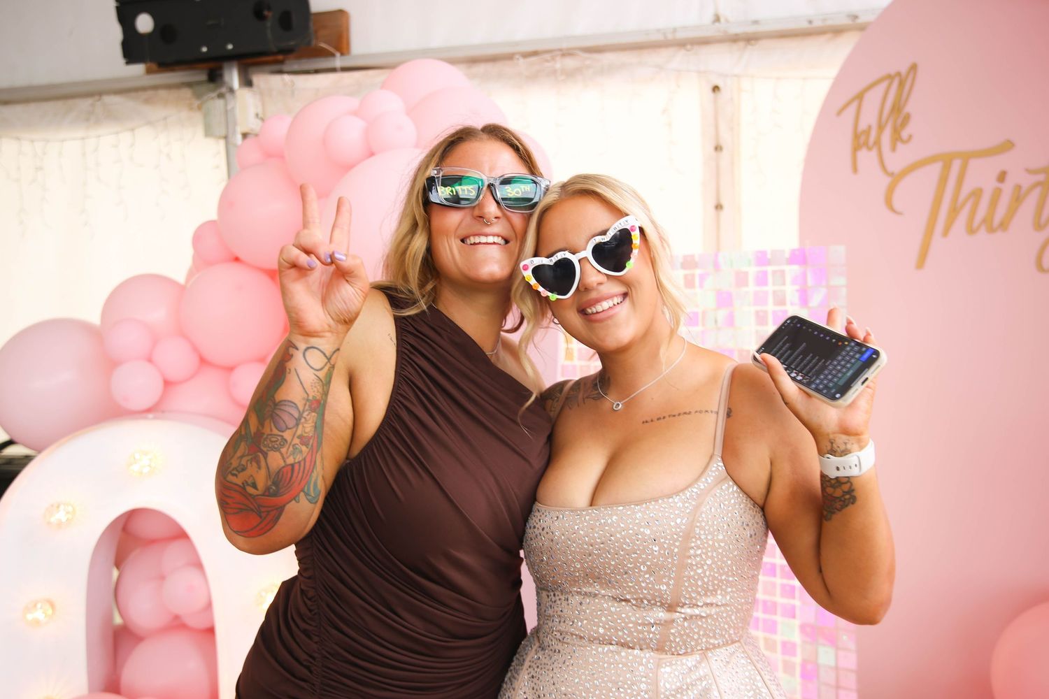 Friends posing with sunglasses against a pink balloon wall at a celebration.