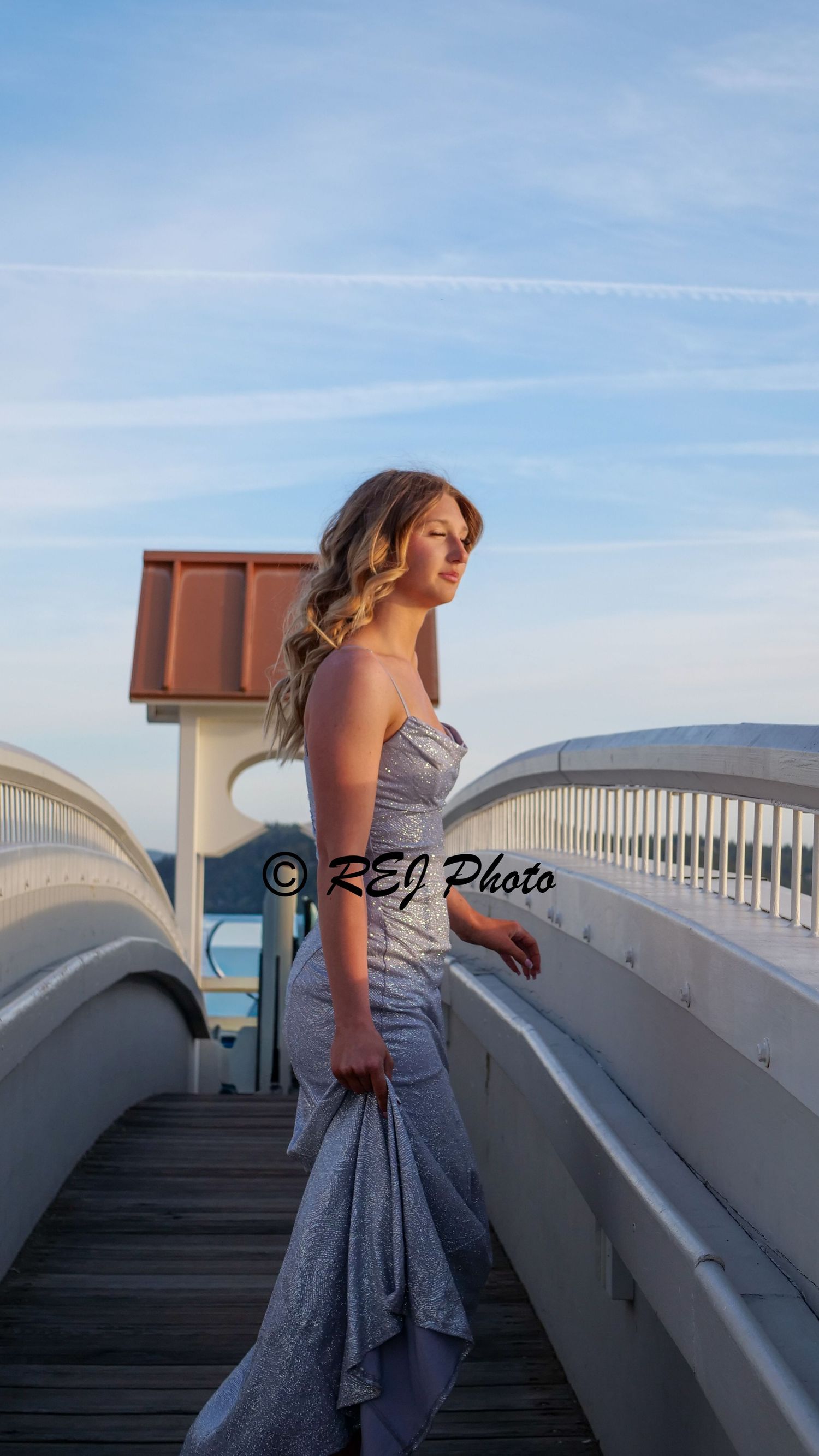 Young woman in silver mermaid gown poses gracefully on a wooden pier bridge under a blue sky at golden hour.