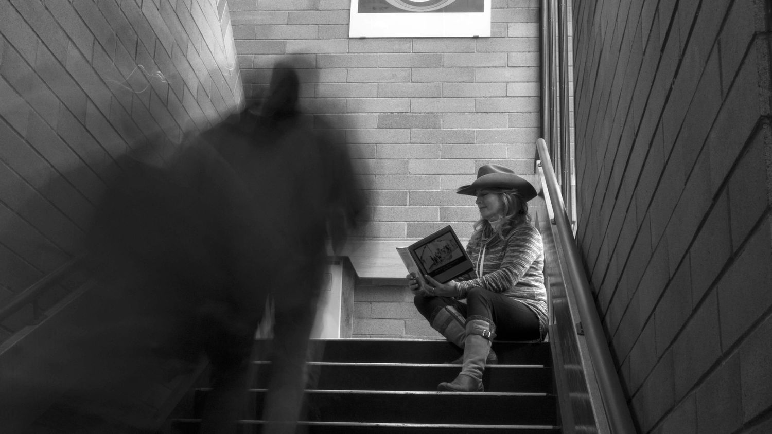 Black and white photo of a man in a cowboy hat reading on stairs while a blurred figure walks past.